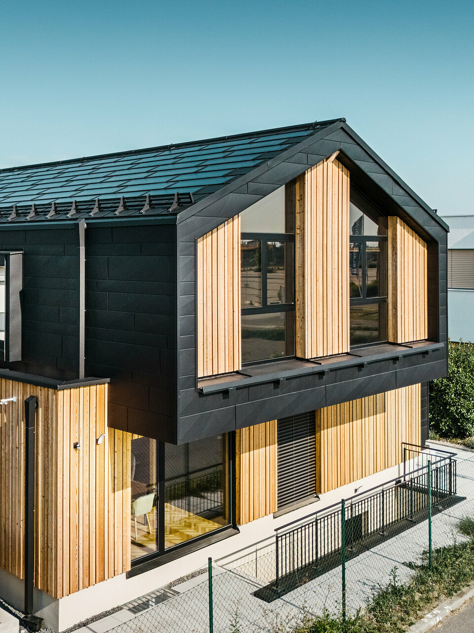 Sprednja stran vzorčne hiše Autark v centru montažnih hiš Blaue Lagune v Vösendorfu (Avstrija). Streha je prekrita s črnimi solarnimi strešnimi ploščami PREFA in strešnimi ploščami R.16 z visoko kakovostnim premazom P.10. Fasada združuje navpične lesene elemente in panele PREFA Siding.X v barvi P.10 črna, kar stavbi daje sodoben in trajnosten značaj. Na desnem robu slike plapola zastava podjetja Erler Bau GmbH.