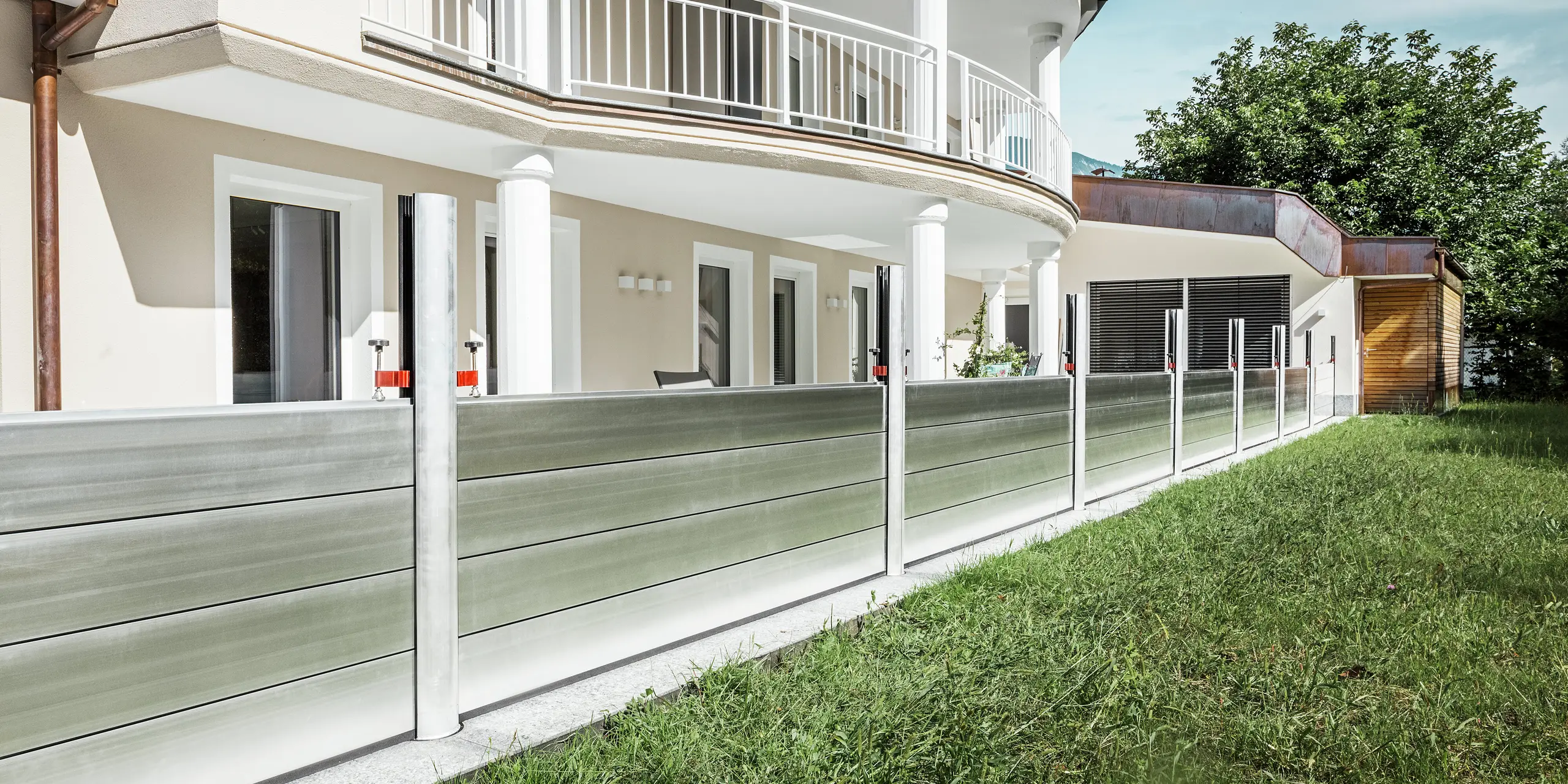 Panoramaansicht eines exklusiven Wohnhauses in Tirol mit installiertem PREFA Hochwasserschutzsystem entlang der Terrasse. Die Dammbalken aus Aluminium verlaufen über die gesamte Gebäudelänge und schützen mehrere Zugänge wie Terrassentüren und Fenster vor möglichem Hochwasser aus dem angrenzenden Gelände. Die moderne Architektur des Hauses mit Rundbalkon und weißen Stützen bildet einen stilvollen Kontrast zum funktionalen Hochwasserschutz aus Aluminium.