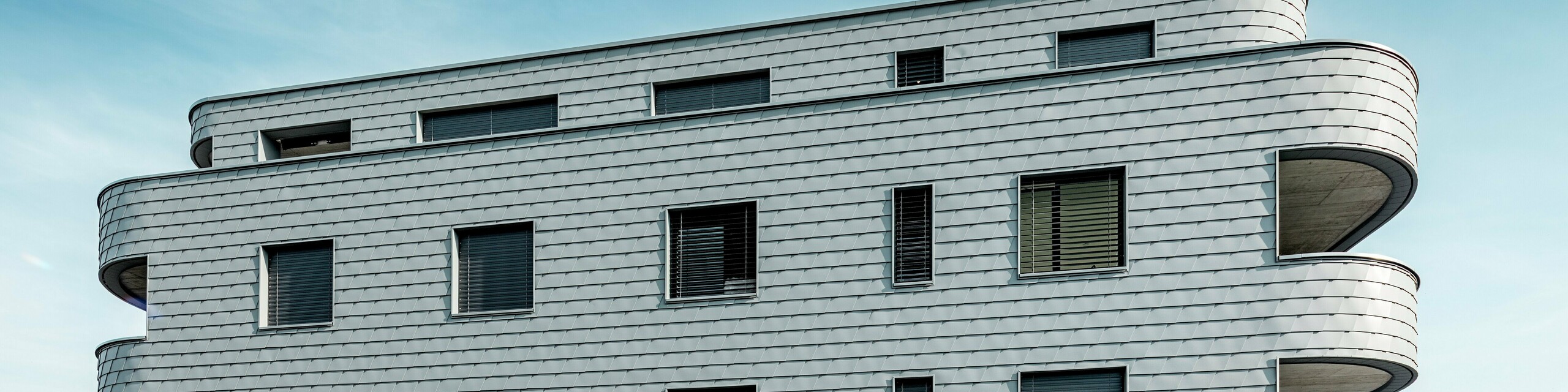 Sprednji pogled na hišo Bernerhaus v Buchsu z nežno zaobljenimi vogali stavbe in neprekinjeno fasado iz PREFA fasadnih skodel v barvi P.10 svetlo siva. Aluminijasta fasada, položena v obliki skodel, ustvarja živahen vzorec in poudarja vodoravne linije ukrivljenih balkonskih pasov. Natančno vgrajene okenske odprtine se harmonično vključujejo v homogeno površino. Zahvaljujoč lahkemu, vremensko odporenemu in trajnemu PREFA aluminiju nastane enostavna za vzdrževanje, oblikovno stabilna ovojnica stavbe z izrazitim videzom pred alpskim ozadjem.