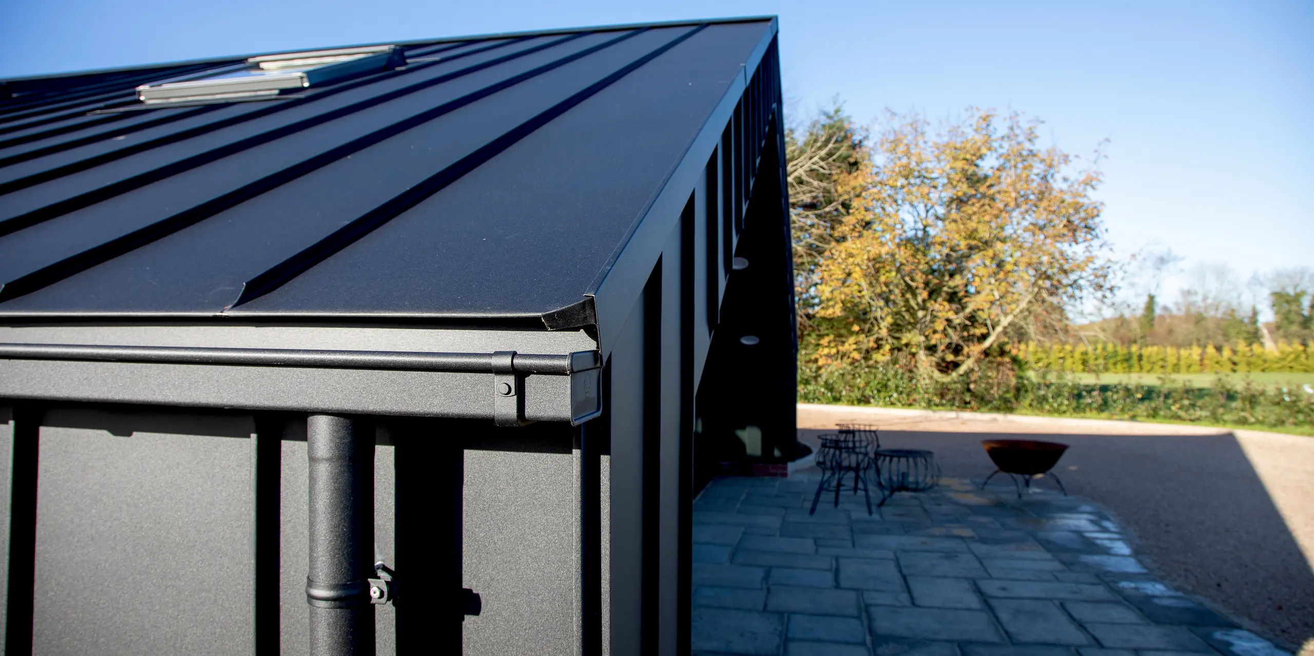 Detailansicht der modernen PREFA Gebäudehülle mit PREFALZ in P.10 Schwarz. Die klaren Stehfalzlinien und die präzise ausgeführte Aluminium-Traufe formen eine markante, elegante Architektur. Die witterungsbeständige Aluminiumoberfläche sorgt für hohe Stabilität und schützt das Freizeitgebäude dauerhaft, während der Außenbereich harmonisch in die moderne Gestaltung übergeht.
