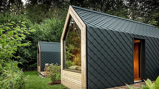 Dve minimalistični hiški Secret Koi Garden stojita v bujni zeleni okolici ob jezeru Natterer See. Fasade in strehe so v celoti obložene z majhnimi PREFA aluminijastimi rombi v barvi P.10 antracit, kar ustvarja izrazito strukturo, podobno ribji koži. Velike steklene površine in topli leseni detajli tvorijo zanimiv kontrast s temnim aluminijem. Trajen in vremensko odporen PREFA aluminij zanesljivo ščiti in poudarja visokokakovostno arhitekturo glampingov.