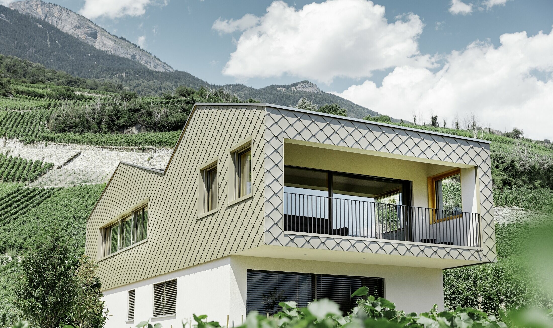 Sodobna enodružinska hiša sredi goric doline Rhône s štirimi različnimi strešnimi površinami in odprto galerijo s fasado iz rombov v bronasti barvi