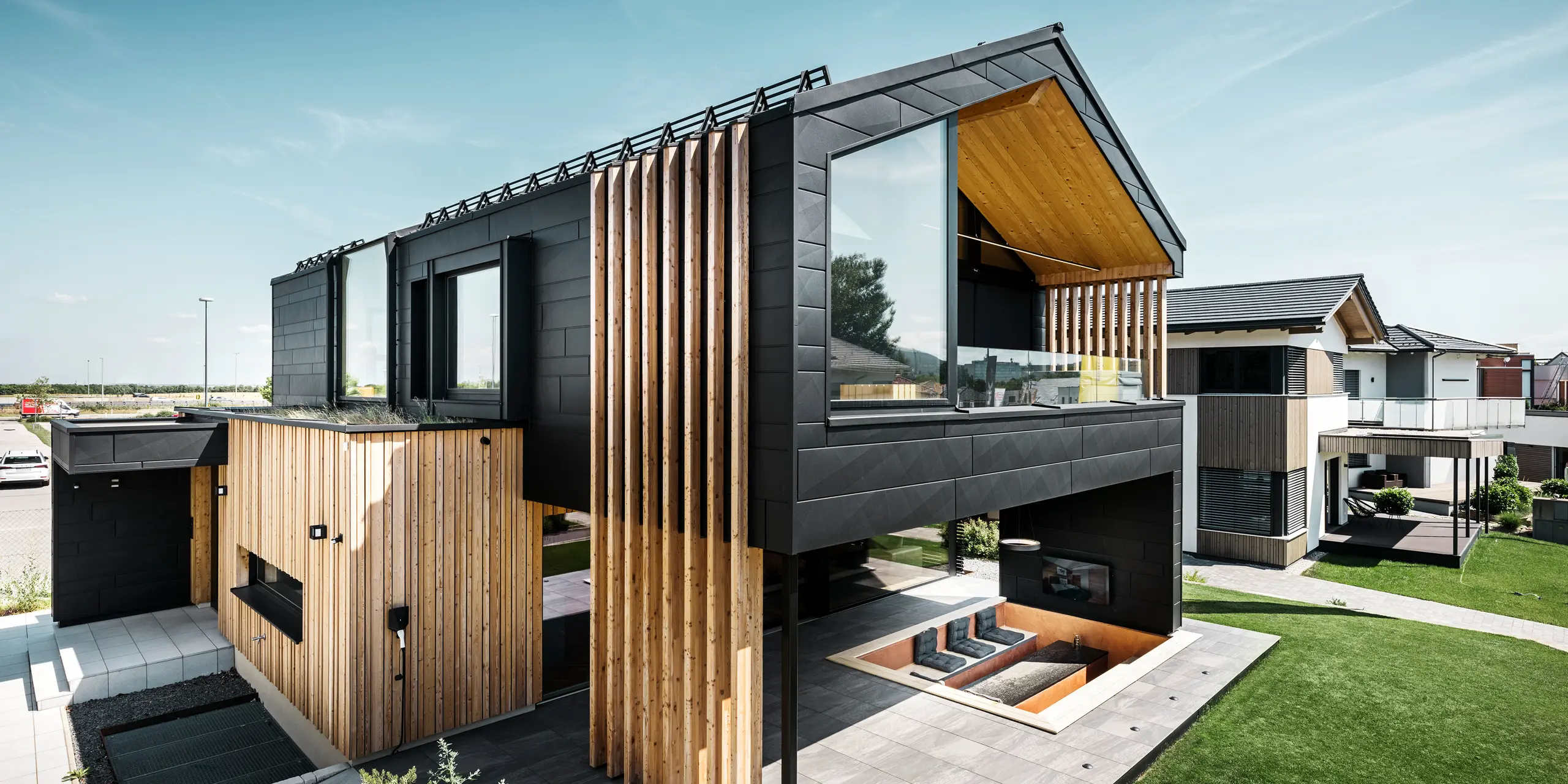 Stranski pogled na vzorčno hišo Autark v Blaue Lagune v Vösendorfu v Avstriji. Zgornje nadstropje navdušuje z velikimi steklenimi površinami in moderno aluminijasto fasado iz elementov PREFA Siding.X v barvi P.10 črna, ki so kombinirani z navpičnimi lesenimi elementi. Prostorna terasa v pritličju z vgrajenim sedežnim kotičkom se brezhibno zliva z arhitekturo. Elegantna in trajnostna zasnova hiše na impresiven način združuje aluminij in les.