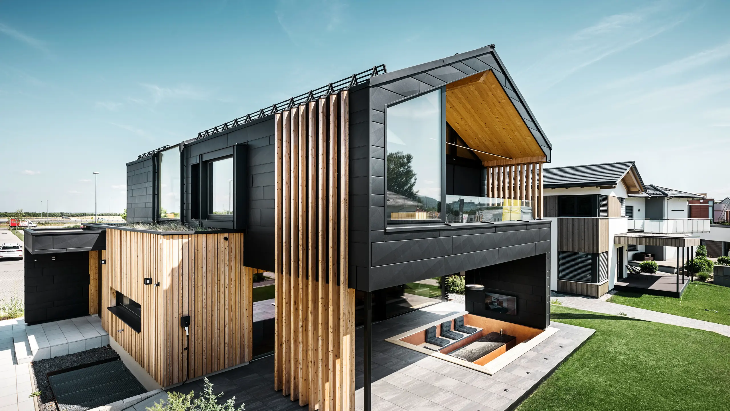 Stranski pogled na vzorčno hišo Autark v Blaue Lagune v Vösendorfu v Avstriji. Zgornje nadstropje navdušuje z velikimi steklenimi površinami in moderno aluminijasto fasado iz elementov PREFA Siding.X v barvi P.10 črna, ki so kombinirani z navpičnimi lesenimi elementi. Prostorna terasa v pritličju z vgrajenim sedežnim kotičkom se brezhibno zliva z arhitekturo. Elegantna in trajnostna zasnova hiše na impresiven način združuje aluminij in les.