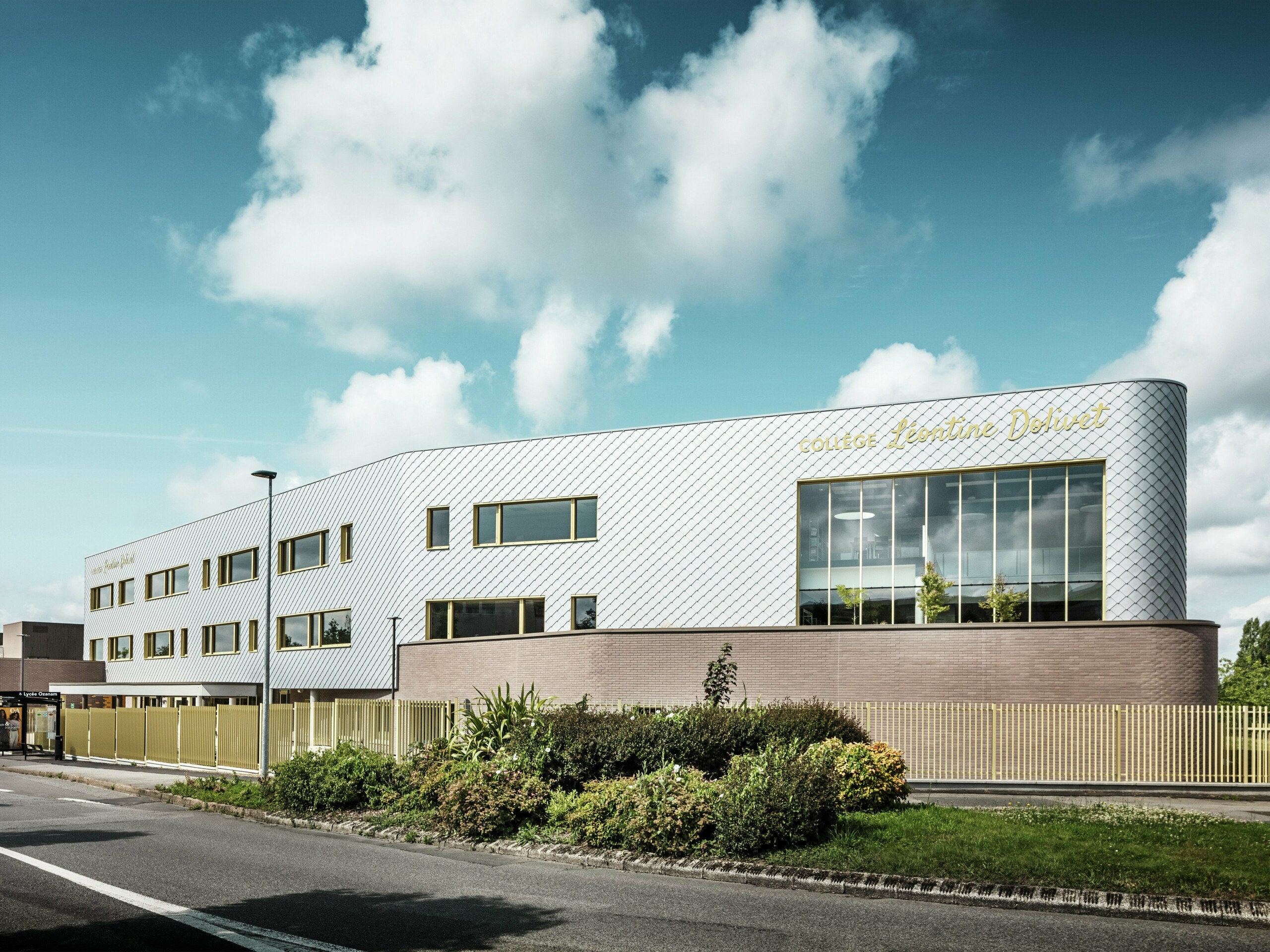 Pogled na Collège Léontine Dolivet z izrazito, srebrno sijočo aluminijasto fasado iz PREFA fasadnih rombov 29 × 29 v kovinsko srebrni barvi. Fasada deluje dinamično zaradi diagonalnega vzorca rombov, medtem ko velike steklene površine in zlata okna ustvarjajo zanimive kontraste. V ospredju so urejene zelene površine in moderna ograja, ki obdajajo stavbo. Trajna fasadna rešitev PREFA združuje eleganco, odpornost proti vremenskim vplivom in trajnost – arhitekturni magnet v Cesson-Sévigné blizu Rennesa.