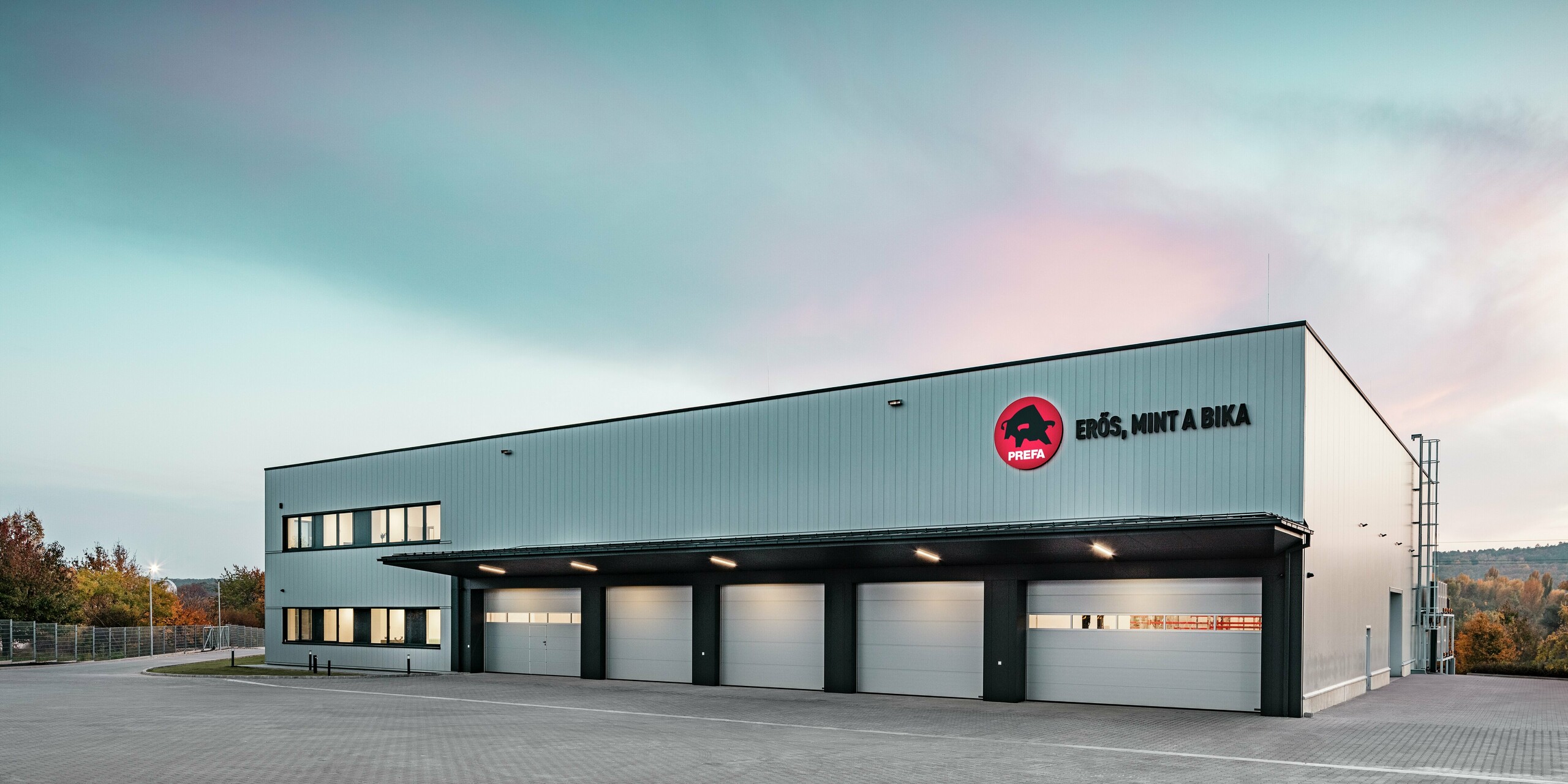 Prostorni skladiščno-logistični objekt PREFA akademije v Törökbálintu (Madžarska) prepriča z moderno in funkcionalno arhitekturo. Fasada s PREFA elementi Siding v srebrno kovinski barvi stavbi daje eleganten, trpežen in vremensko odporen videz. Prepoznavni logotip PREFA s sloganom »Erős, mint a bika« (»Močan kot bik«) poudarja kakovost in robustnost aluminijastih izdelkov.