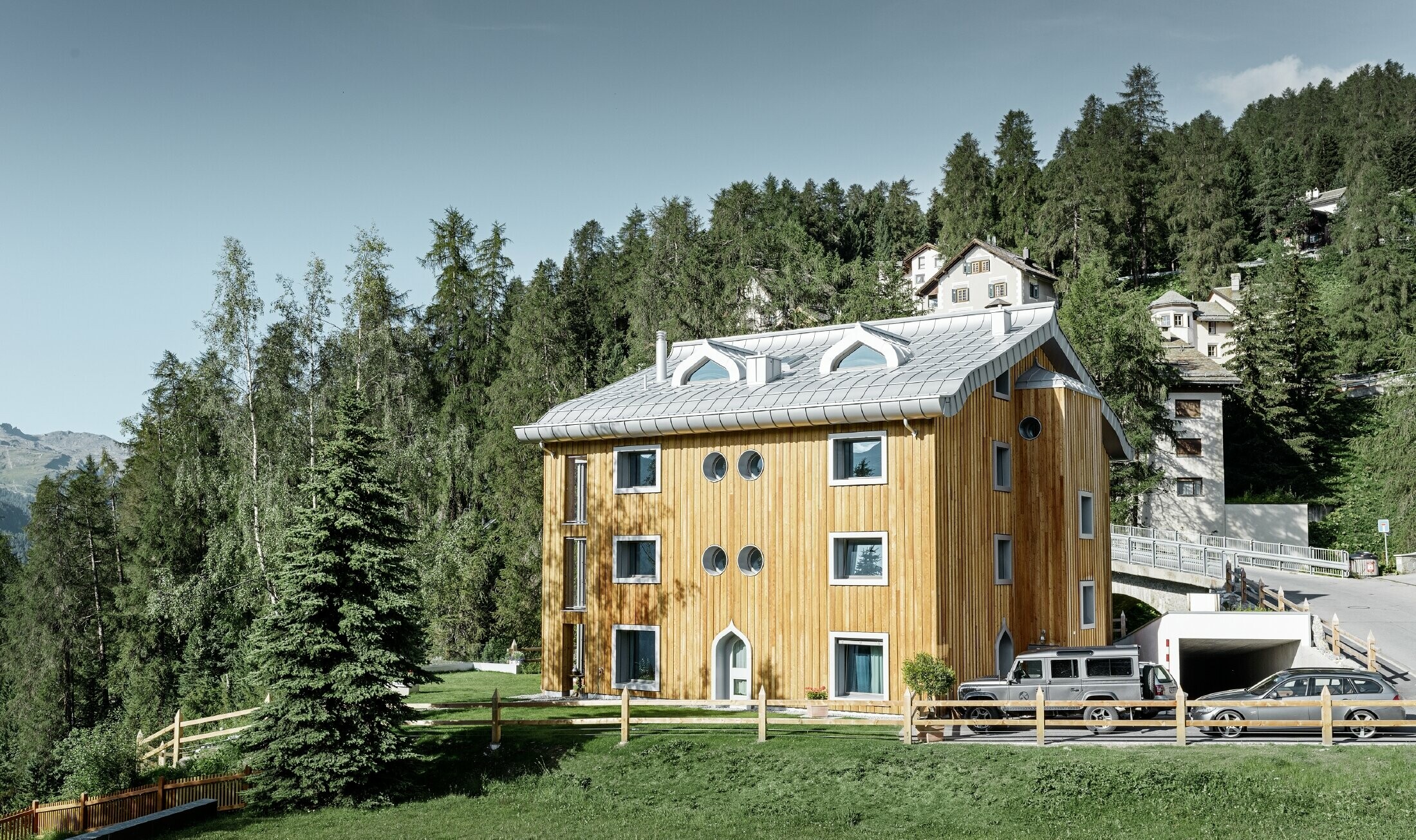 Stanovanjski kompleks v St. Moritzu z leseno fasado in aluminijasto streho z zavihanim kapom v kovinsko srebrni barvi