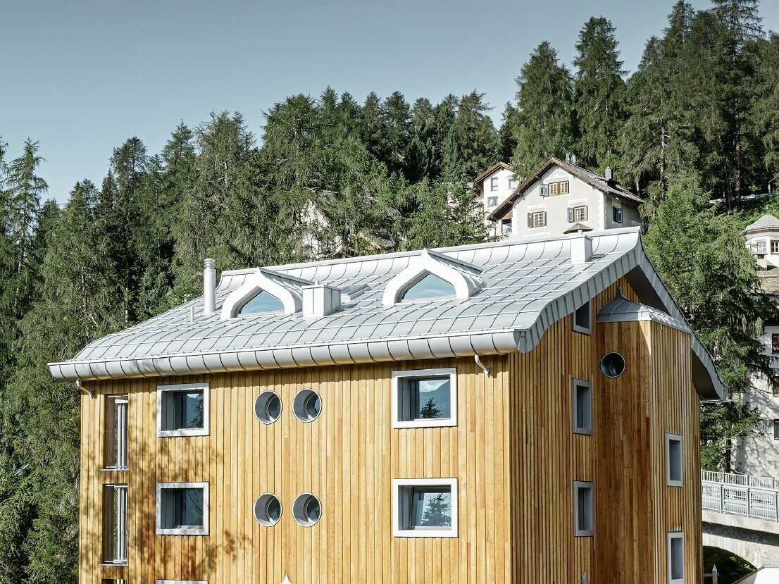 Stanovanjski kompleks v St. Moritzu z leseno fasado in aluminijasto streho z zavihanim kapom v kovinsko srebrni barvi