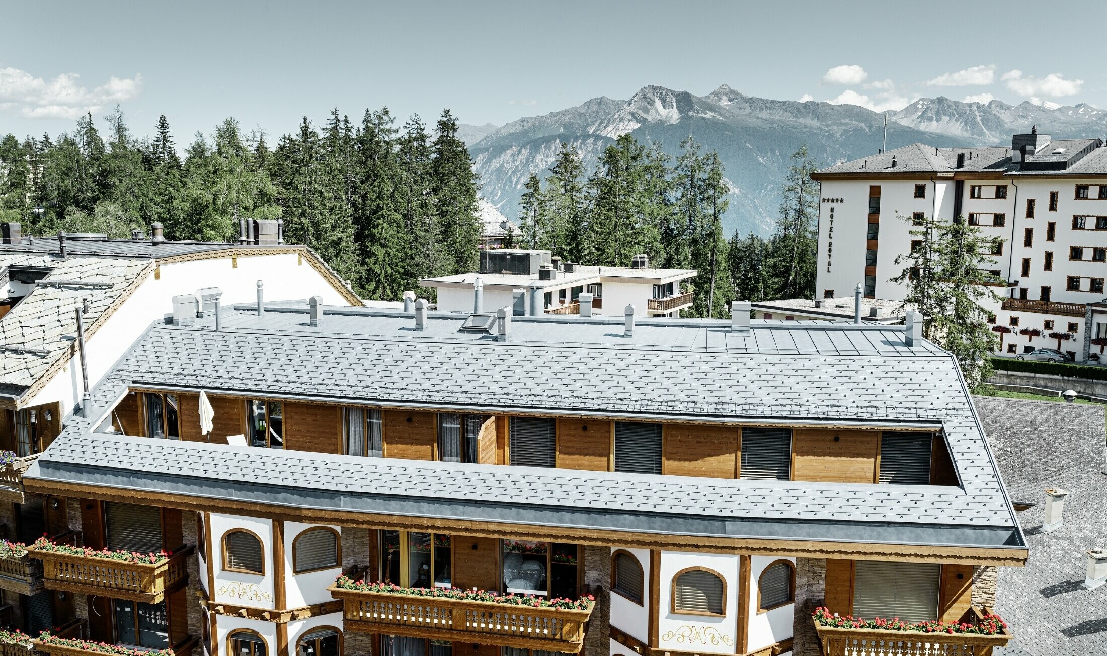 Večdružinska hiša v Crans-Montani z gorami v ozadju in fasado z igrivimi lesenimi elementi in streho iz aluminijastih strešnih skodel PREFA v kamnito sivi barvi