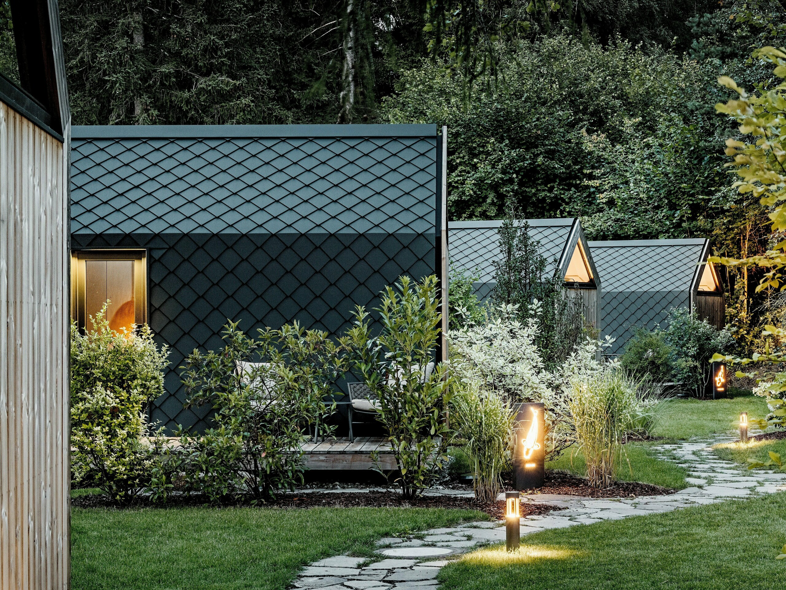 Osvetljena vrtna pot vodi skozi glamping resorta Secret Koi Garden ob jezeru Natterer see do več minimalističnih majhnih hišic. Kompaktne stavbe so v celoti obložene s strešnimi in fasadnimi rombi PREFA iz aluminija v barvi P.10 antracit, katerih struktura, podobna ribji koži, v večerni svetlobi deluje elegantno. Obdane z bujno zeleno naravo, trajne in vremensko odporne PREFA aluminijaste obloge združujejo visokokakovostno arhitekturo z naravo, umirjenostjo in trajnostnim oblikovanjem.