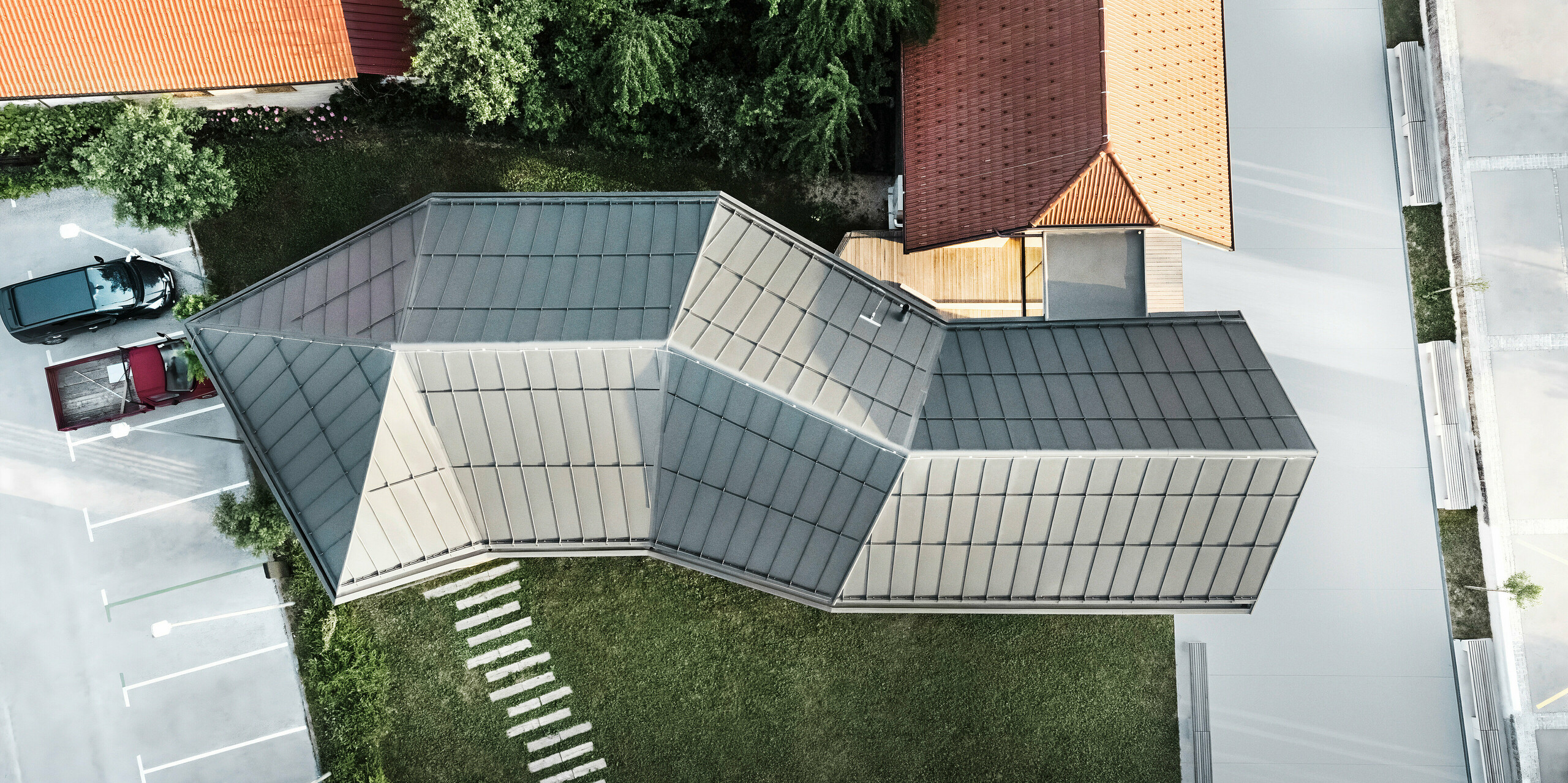 Pogled iz ptičje perspektive na turistično informacijski center za obiskovalce na Igu v Sloveniji, ki navdušuje s kompleksno geometrijsko streho s sistemom PREFALZ v P.10 temno sivi barvi. Izstopajoča strešna konstrukcija odraža inovativno arhitekturo in prilagajanje raznoliki okolici s sosednjimi tradicionalnimi strehami iz strešnikov. Natančna izdelava in sodoben dizajn poudarjata strokovno znanje PREFA na področju  sodobnega oblikovanja streh.