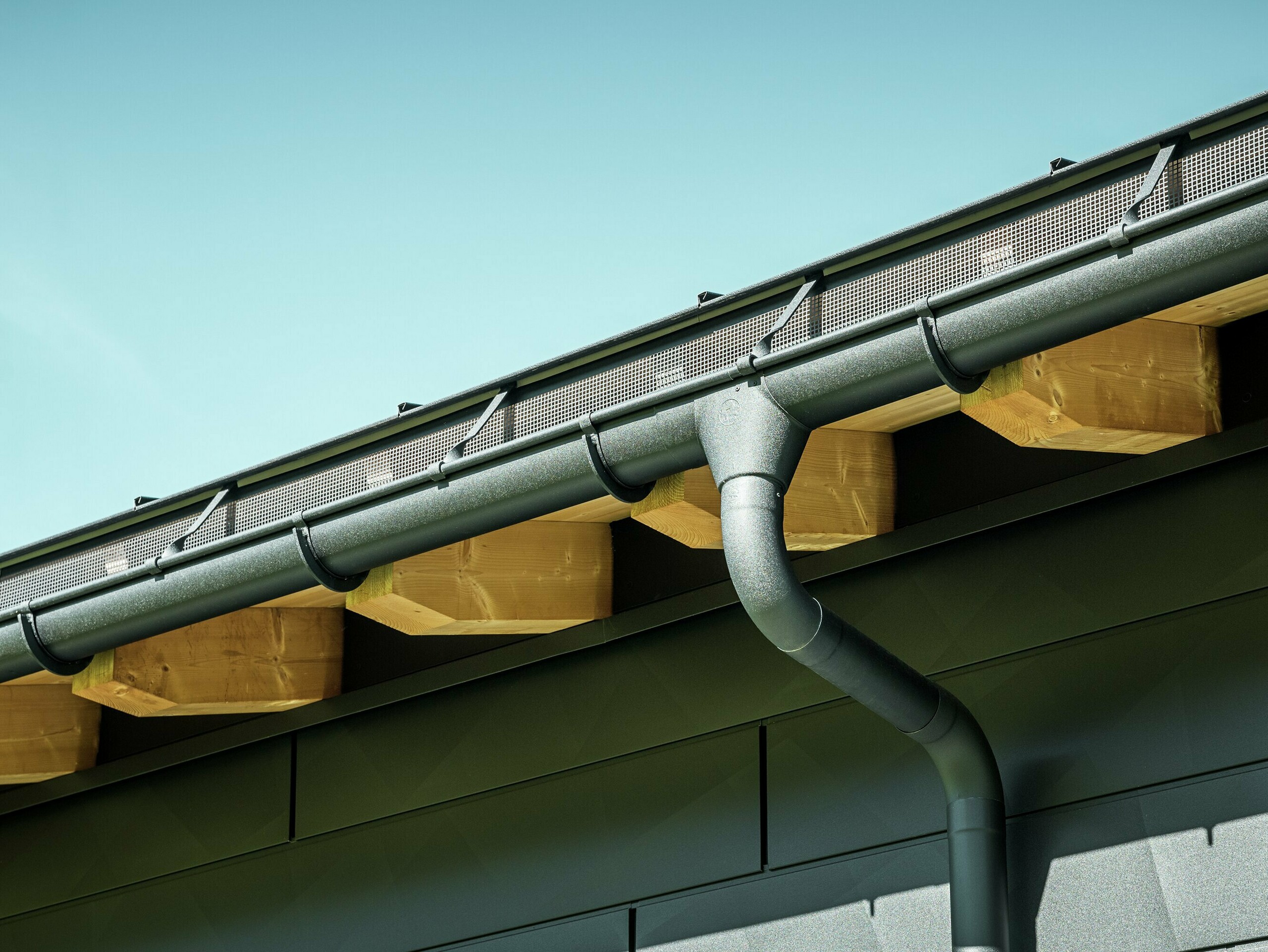 Približek fasade moderne enodružinske hiše v Ascharini v Švici. Strani stavbe so obložene z izrazitimi fasadnimi paneli PREFA Siding.X v barvi P.10 črna. Barvno usklajeno je tudi odvodnjavanje strehe PREFA s strešnim žlebom in iztočno cevjo.