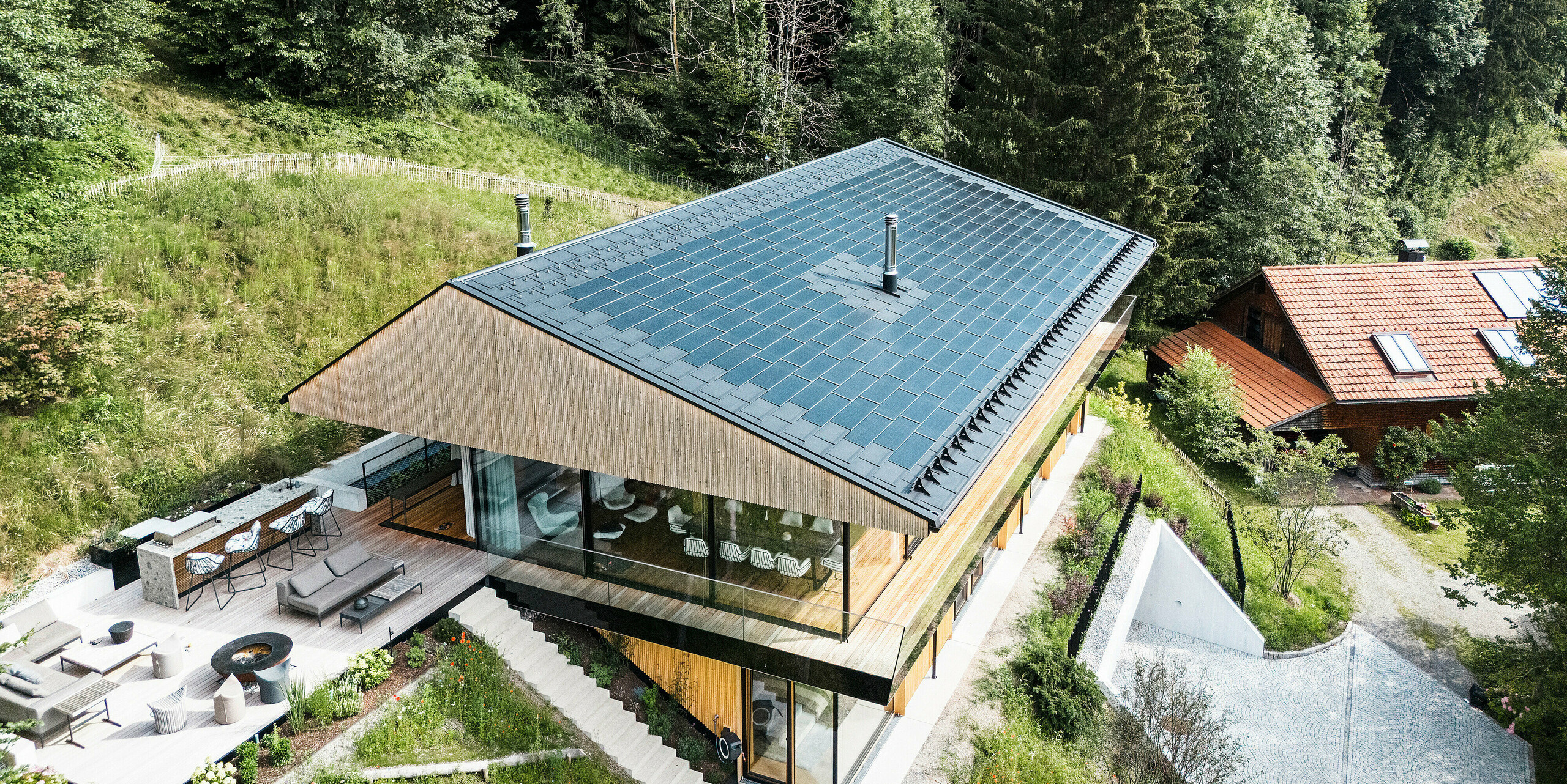 Pogled na kakovostno zunanjo kuhinjo enodružinske hiše v kraju Egg v Vorarlbergu. Sodobna stavba je bila pokrita z visokokakovostnimi solarnimi strešnimi ploščami PREFA in strešnimi ploščami R.16 v črni barvi P.10. Aluminijasta streha z vgrajenim fotovoltaičnim sistemom omogoča trajnostno pridobivanje energije ter se ujema z vabljivo leseno fasado in okoliško naravo.