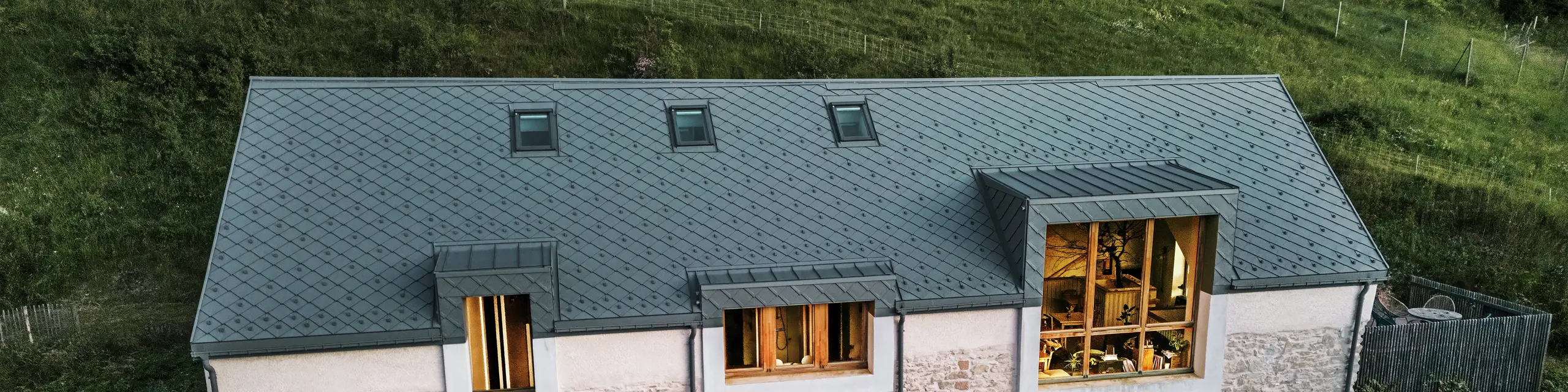 Večerni pogled na obnovljeni skedenj „La Grange à Gaby“ v Enginsu v Franciji. Stavba združuje tradicionalno kamnito zidovje s sodobno aluminijasto streho PREFA, izdelano iz rombov 29 × 29 cm. Velika okna in osvetljena notranjost poudarjajo vabljivo vzdušje novega kulturnega centra.