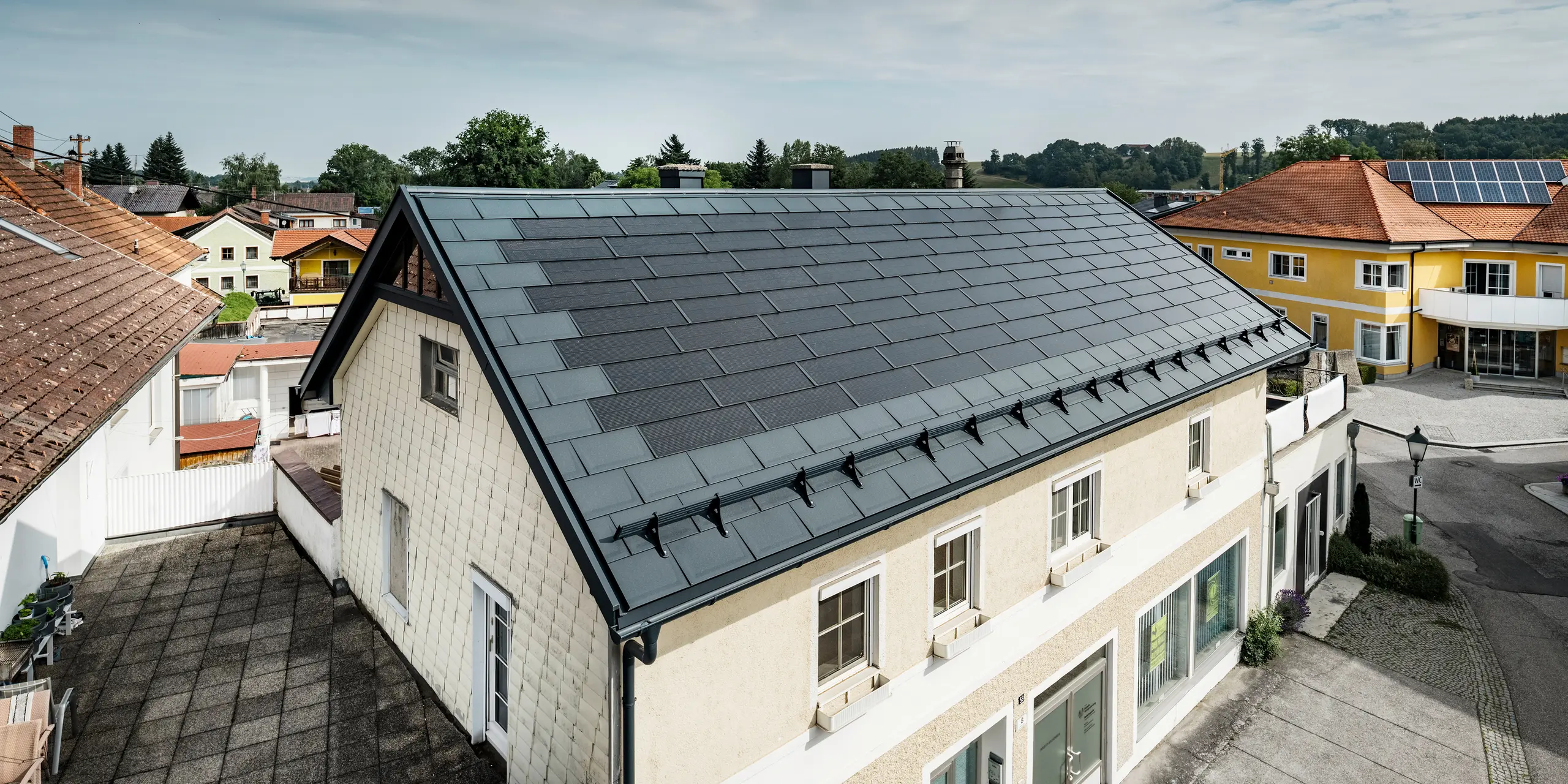 Pogled od zgoraj na enodružinsko hišo v Mettmachu v Avstriji. Starejši objekt je bil prenovljen s PREFA solarnimi strešnimi ploščami v antracitni barvi. Sistemi za zaščito pred snegom in jasne linije solarnih strešnih plošč poudarjajo čisto izdelavo aluminijastih izdelkov PREFA. V ozadju je v kontrastu s streho sosednje stavbe, ki je obarvana oranžno. Fotonapetostni moduli so nameščeni na strehi sosednje enodružinske hiše v klasični strešni rešitvi, kar nakazuje na pestrost energijsko učinkovite izvedbe streh.