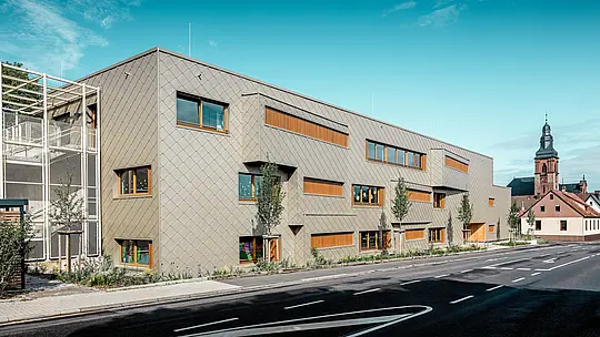 V jasni dnevni svetlobi se sodobni vrtec v mestu Alzenau s svojo izrazito PREFA aluminijasto fasado iz bronastih fasadnih rombov 44 × 44 kaže v vsej svoji jasnosti. Enotna struktura lusk, prekinjena z zamaknjenimi okni in navpičnimi lesenimi lamelami, fasadi daje globino in ritem. Na levi strani slike je vidno filigransko stopnišče s kovinsko mrežasto strukturo, ki zagotavlja preglednost in lahkotnost. V ozadju stoji poznogotska cerkev v Alzenau – simbol harmonične povezave zgodovinske gradbene kulture in sodobne arhitekture. Brez vzdrževanja, trajen PREFA fasadni sistem podpira trajnostno, funkcionalno in estetsko visokokakovostno oblikovanje te napredne izobraževalne stavbe.