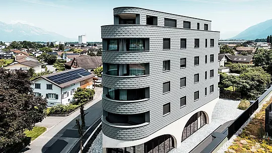 Letalski posnetek stavbe Bernerhaus v Buchsu z značilno trikotno obliko in nežno zaobljenimi balkoni. Fasada iz PREFA fasadnih skodel v barvi P.10 svetlo siva obdaja stavbo kot luskasta aluminijasta koža in poskrbi za živahno igro svetlobe in sence. Nad masivnim betonskim podstavkom z elegantnimi arkadami deluje lahka, vremensko odporna PREFA aluminijasta fasada še posebej natančna, trajna in enostavna za vzdrževanje – sodoben magnet za poglede sredi alpskega mestnega okolja.