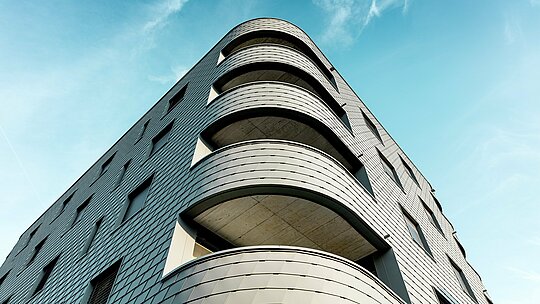 Dinamičen pogled od spodaj na moderno poslovno-stanovanjsko stavbo z izrazito zaobljenim vogalom in tekočimi balkonskimi pasovi. Fasada v obliki skodel iz PREFA fasadnih skodel v barvi P.10 svetlo siva se kot natančno obdelana aluminijasta koža ovije okoli stavbe in poudari mehke zaobljenosti. Enotna površina poudarja jasne linije, medtem ko lahki, vremensko odporni PREFA aluminij tudi pri močnih krivinah zagotavlja najvišjo oblikovno stabilnost, dolgo življenjsko dobo in minimalno vzdrževanje – arhitekturni magnet za poglede pred sijočim modrim nebom.