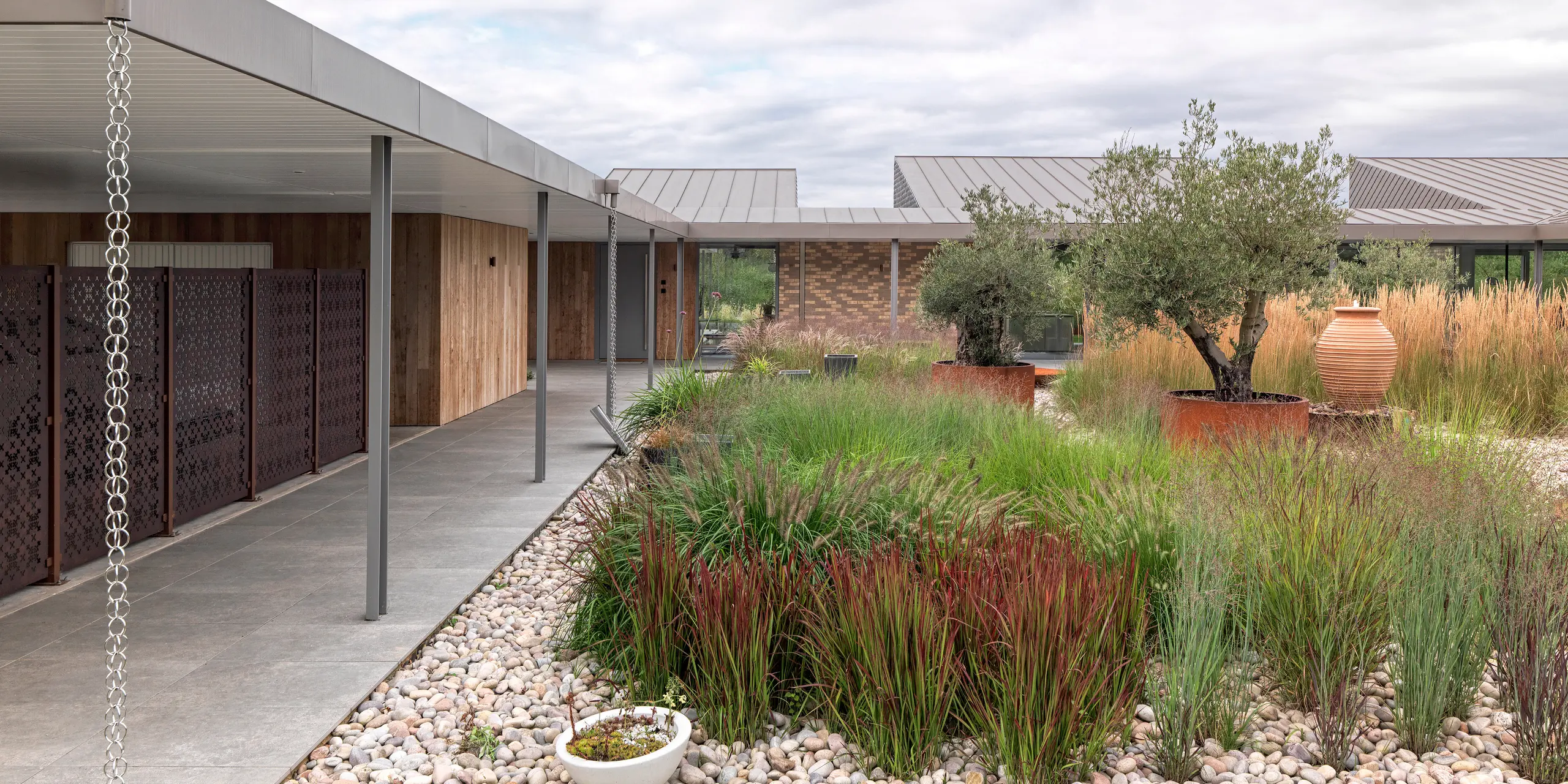 Detailansicht des modernen Wohnhauses in West Sussex mit Flachdach aus PREFALZ Aluminium in P.10 Bronze. Zu sehen ist die überdachte, klar strukturierte Terrassenanlage mit filigranen Säulen, stilvoll integrierten Regenablaufketten und einer abwechslungsreichen Gartengestaltung mit Ziergräsern, Olivenbäumen und Natursteinen. Die langlebigen PREFA Dachprodukte aus Aluminium unterstreichen die moderne Linienführung und fügen sich harmonisch in das nachhaltige Architekturkonzept ein.