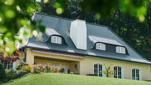 Enodružinska hiša s sanirano streho s strešnimi skodlami PREFA v antracitni barvi z zaobljenimi mansardnimi okni in belim dimnikom.