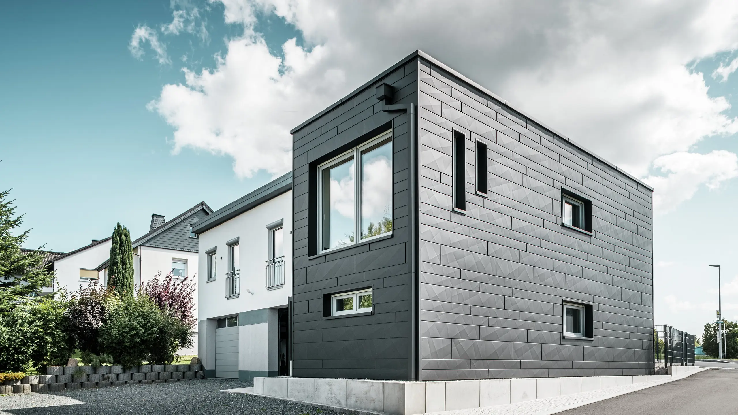 Elegantna in moderna fasada obnovljene enodružinske hiše v Teuschnitzu je obložena z jasno strukturiranimi ploščami PREFA Siding.X v barvi P.10 črna. Geometrična razporeditev visokokakovostnih pločevinastih plošč daje stavbi izrazito, brezčasno estetiko. Okna so obrobljena s črnimi aluminijastimi kompozitnimi ploščami, kar harmonično dopolnjuje celoten dizajn. Vgrajen PREFA sistem za odvodnjavanje strehe s pravokotno iztočno cevjo zagotavlja funkcionalno in estetsko rešitev.