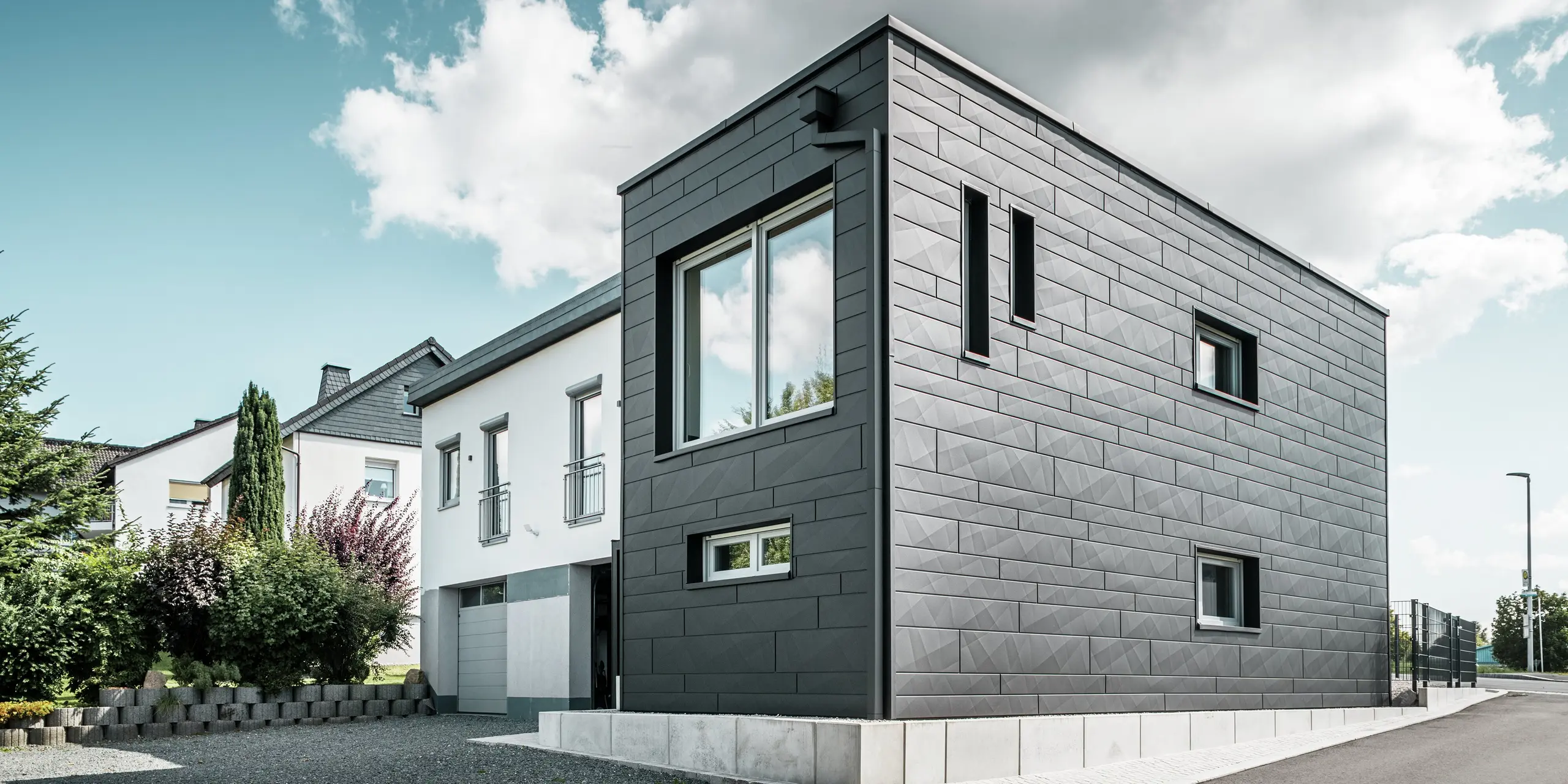 Elegantna in moderna fasada obnovljene enodružinske hiše v Teuschnitzu je obložena z jasno strukturiranimi ploščami PREFA Siding.X v barvi P.10 črna. Geometrična razporeditev visokokakovostnih pločevinastih plošč daje stavbi izrazito, brezčasno estetiko. Okna so obrobljena s črnimi aluminijastimi kompozitnimi ploščami, kar harmonično dopolnjuje celoten dizajn. Vgrajen PREFA sistem za odvodnjavanje strehe s pravokotno iztočno cevjo zagotavlja funkcionalno in estetsko rešitev.