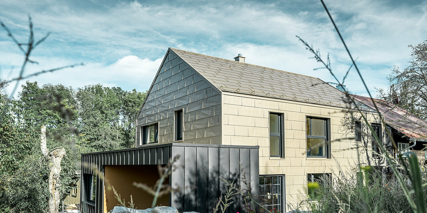 Na stanovanjski hiši brez napušča sta streha in fasada oblečeni v aluminijasti panel FX.12 PREFA v bronasti barvi. Tik ob njej stoji garaža z ravno streho, ki ima fasado s kotnim stoječim kleparskim spojem.
