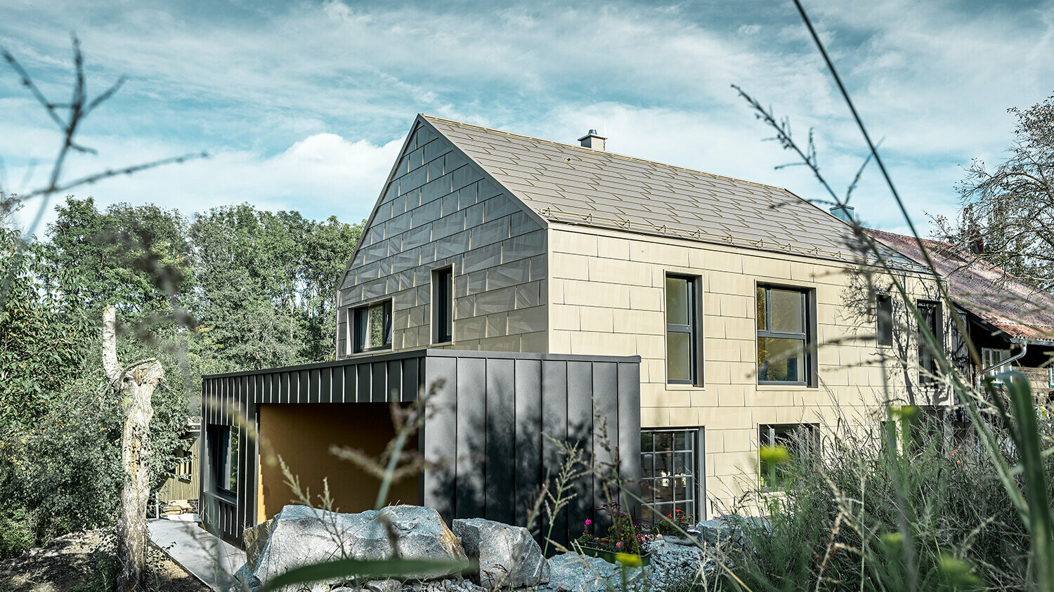 Na stanovanjski hiši brez napušča sta streha in fasada oblečeni v aluminijasti panel FX.12 PREFA v bronasti barvi. Tik ob njej stoji garaža z ravno streho, ki ima fasado s kotnim stoječim kleparskim spojem.