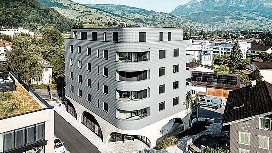 Letalski posnetek hiše Bernerhaus v Buchsu z značilno trikotno obliko stavbe in nežno zaobljenimi balkoni sredi alpske pokrajine. Neprekinjena fasada iz PREFA fasadnih  skodel v barvi P.10 svetlo siva obdaja stavbo kot luskasta aluminijasta koža in ustvarja živahno igro svetlobe in strukture. Nad masivnim betonskim podstavkom z arkadami deluje lahka, vremensko odporna PREFA aluminijasta fasada še posebej natančna, trajna in enostavna za vzdrževanje – sodoben magnet za poglede v mestni podobi.