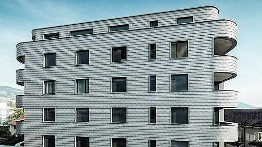Sprednji pogled na hišo Bernerhaus v Buchsu z nežno zaobljenimi vogali stavbe in neprekinjeno fasado iz PREFA fasadnih skodel v barvi P.10 svetlo siva. Aluminijasta fasada, položena v obliki skodel, ustvarja živahen vzorec in poudarja vodoravne linije ukrivljenih balkonskih pasov. Natančno vgrajene okenske odprtine se harmonično vključujejo v homogeno površino. Zahvaljujoč lahkemu, vremensko odporenemu in trajnemu PREFA aluminiju nastane enostavna za vzdrževanje, oblikovno stabilna ovojnica stavbe z izrazitim videzom pred alpskim ozadjem.
