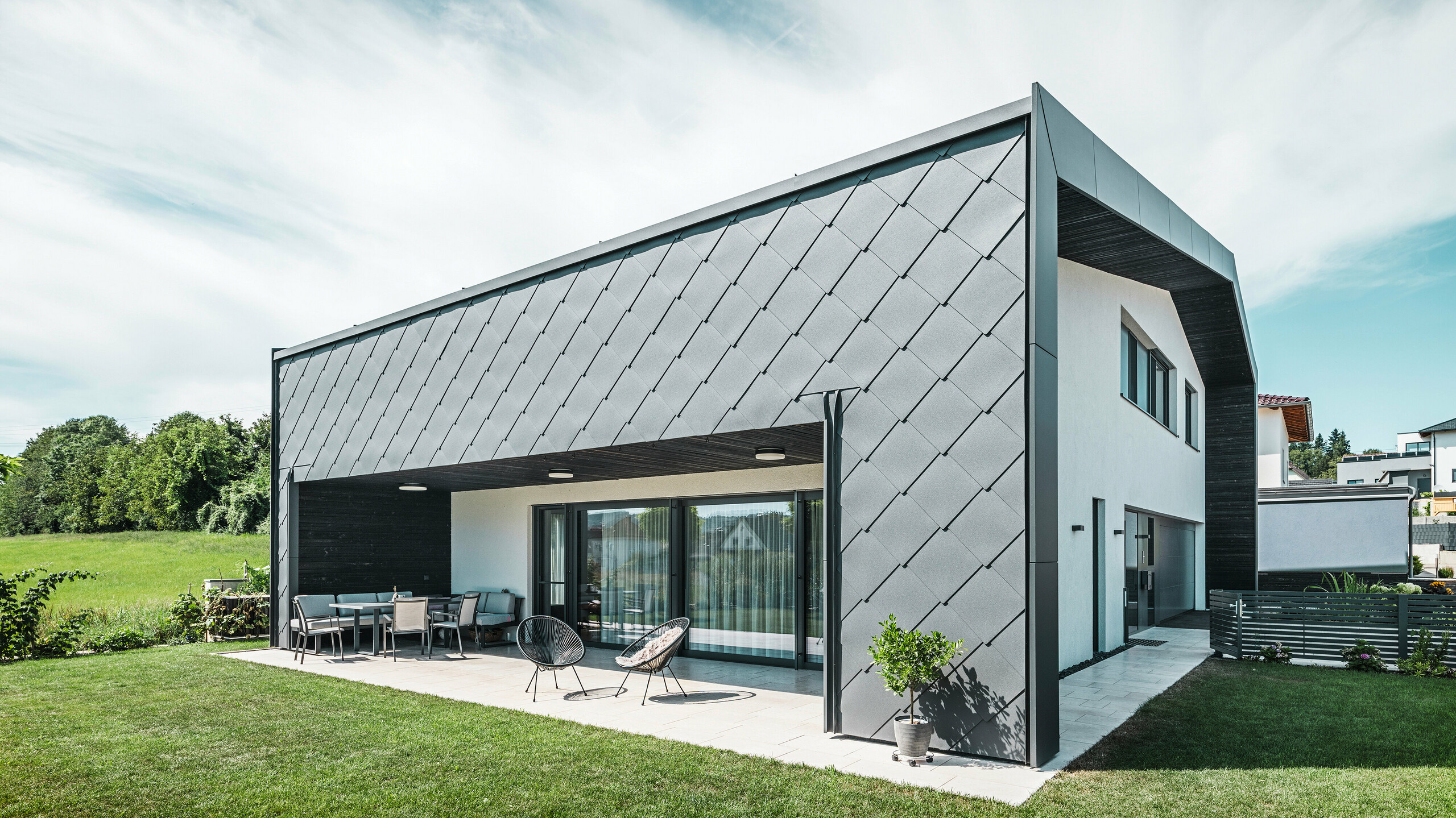 Urejena vrtna površina enodružinske hiše v Aschachu. Stavba je stilsko pokrita s temno sivimi fasadnimi rombi PREFA iz aluminija 44 × 44. Pokrita terasa ponuja prijeten prostor za sedenje z razgledom na zeleno travnato površino. Elegantna aluminijeva obloga v kombinaciji z belimi ometnimi površinami zagotavlja moderno in vremensko odporno arhitekturo. Velika okna prepuščajo veliko dnevne svetlobe v notranjost in ustvarjajo odprto, svetlo vzdušje.