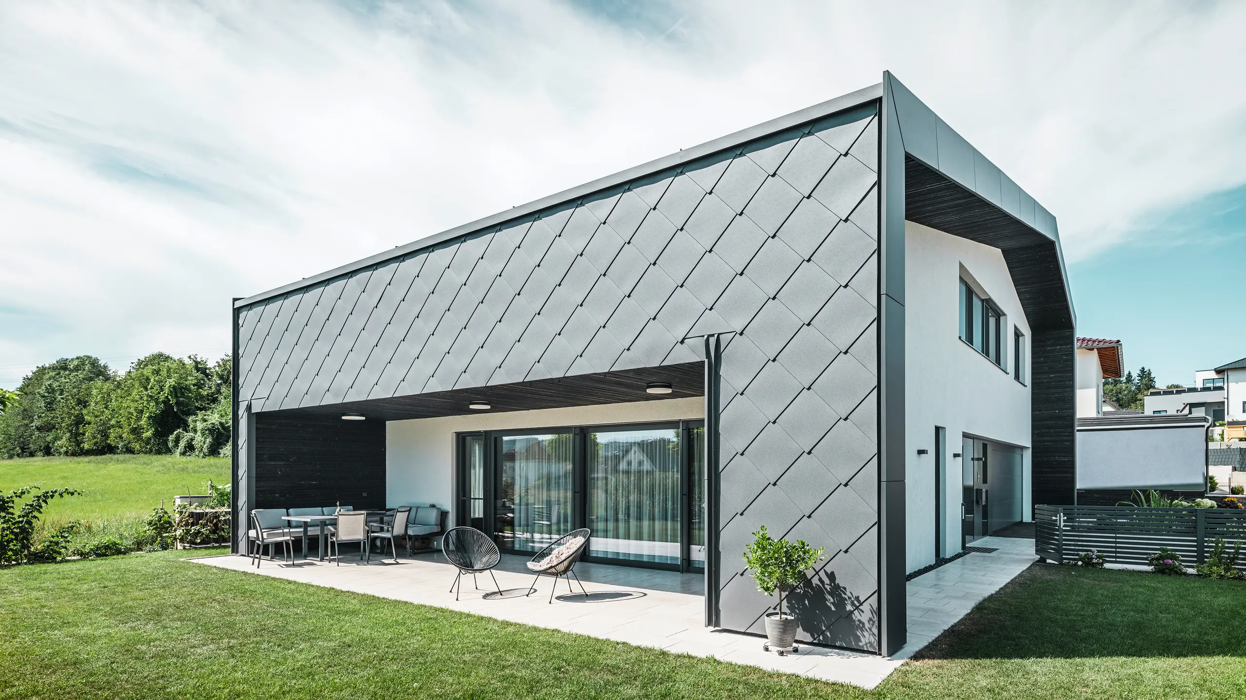 Urejena vrtna površina enodružinske hiše v Aschachu. Stavba je stilsko pokrita s temno sivimi fasadnimi rombi PREFA iz aluminija 44 × 44. Pokrita terasa ponuja prijeten prostor za sedenje z razgledom na zeleno travnato površino. Elegantna aluminijeva obloga v kombinaciji z belimi ometnimi površinami zagotavlja moderno in vremensko odporno arhitekturo. Velika okna prepuščajo veliko dnevne svetlobe v notranjost in ustvarjajo odprto, svetlo vzdušje.