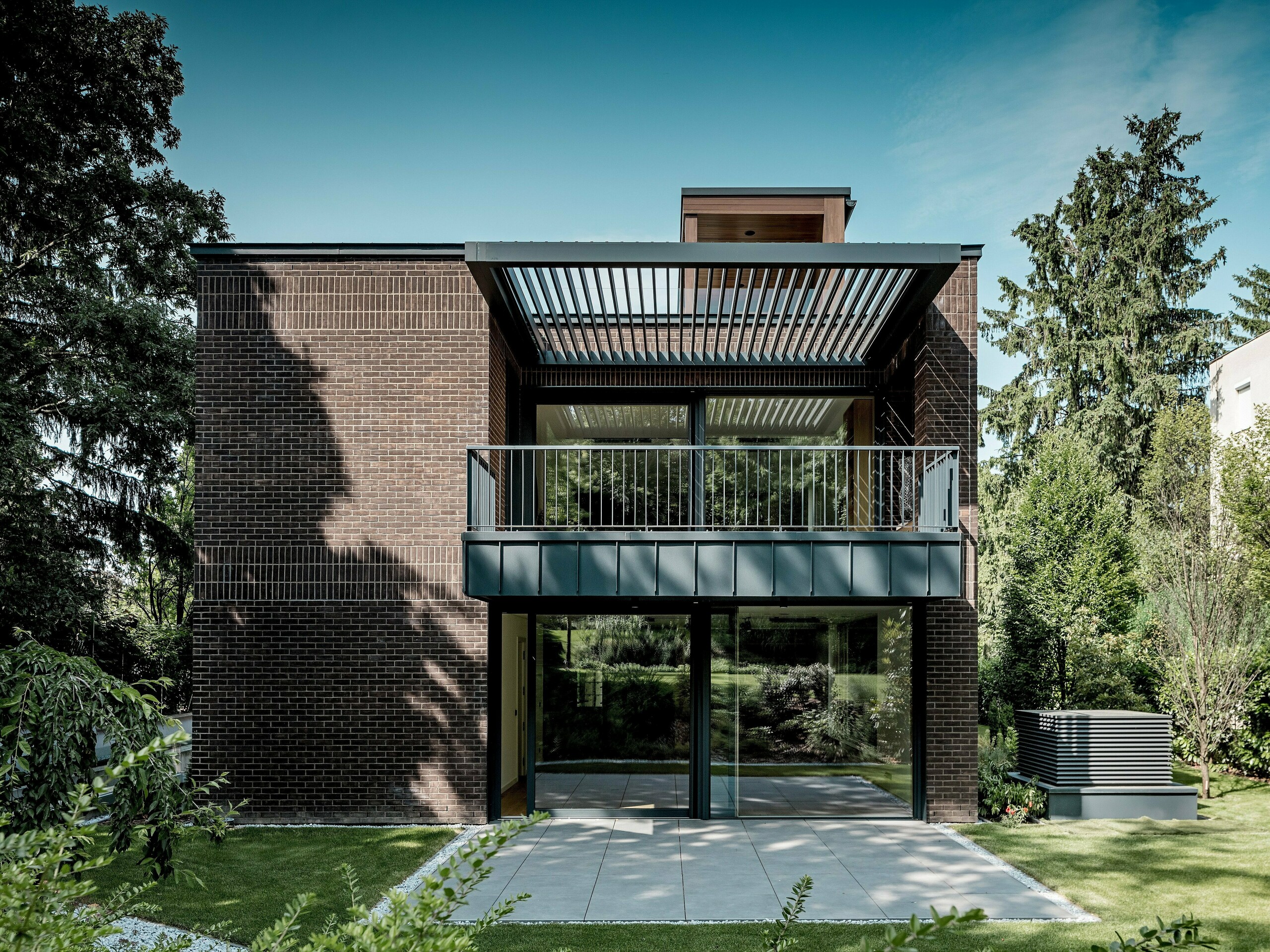 Fotografija prikazuje stran moderne večgeneracijske vile v Budimpešti, obrnjeno proti vrtu, z jasno strukturirano fasado iz temno rdečega klinkerja in PREFALZ aluminija v barvi P.10 antracit. Veliki balkon s filigransko kovinsko konstrukcijo in nad njim nameščenimi lamelami ustvarja zanimivo igro svetlobe in sence. Steklene fasade od tal do stropa odpirajo notranji prostor proti zeleni vrtni pokrajini. Natančno izvedeni aluminijasti detajli poudarjajo trajnost, vremensko odpornost in arhitekturno jasnost rešitev PREFA, ki stavbi dajejo izrazit, brezčasen videz.