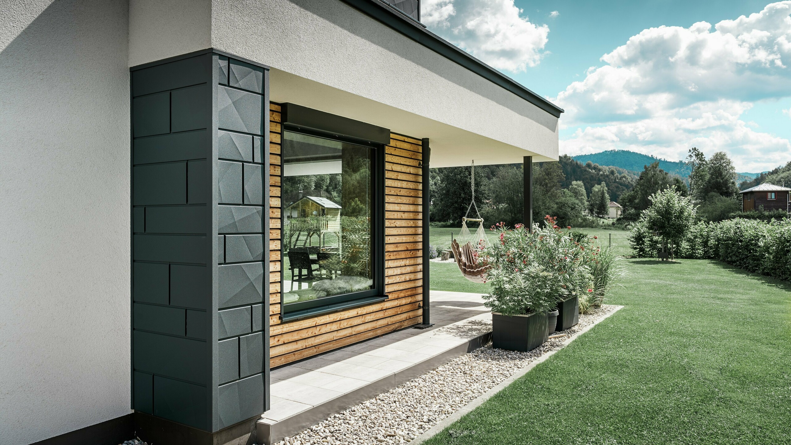 Delni pogled na novo zgrajeno samostojno hišo. Na fasadi so bili uporabljeni različni materiali. Bela ometana fasada je poudarjena z aluminijastimi elementi PREFA Siding.X v antracitni barvi in vodoravnimi lesenimi paneli, ki obdajajo veliko okno. Na terasi visi viseči stol.