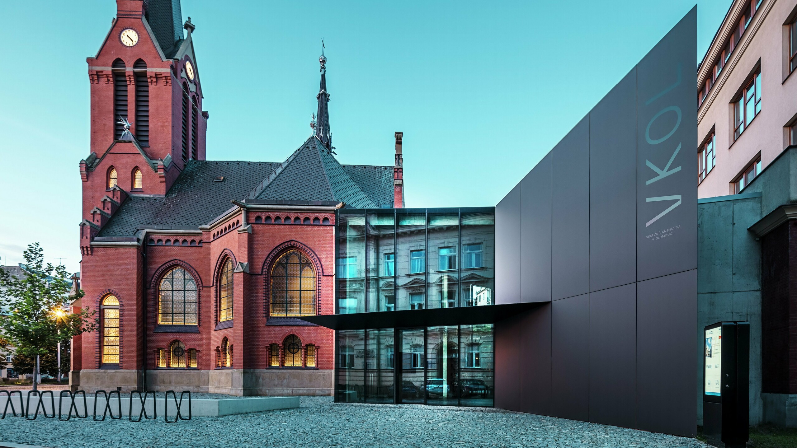 Slika prikazuje impresivno prepletanje zgodovinske Rdeče cerkve in moderne nove stavbe v Olmützu. Cerkev s svojimi značilnimi gotskimi okni in izrazitim stolpom tvori zgodovinsko hrbtenico kompleksa, medtem ko moderni prizidek s svojo elegantno črno fasado iz aluminijastih kompozitnih plošč PREFABOND predstavlja sodoben arhitekturni odgovor. Velike steklene površine omogočajo pogled v notranjost nove stavbe, kjer okolica z odsevi v oknih odraža zanimivo interakcijo med starim in novim. Natančna obdelava plošč PREFABOND poudarja sodobno estetiko, medtem ko harmonična integracija v zgodovinsko okolje navdušuje tako funkcionalno kot vizualno. 