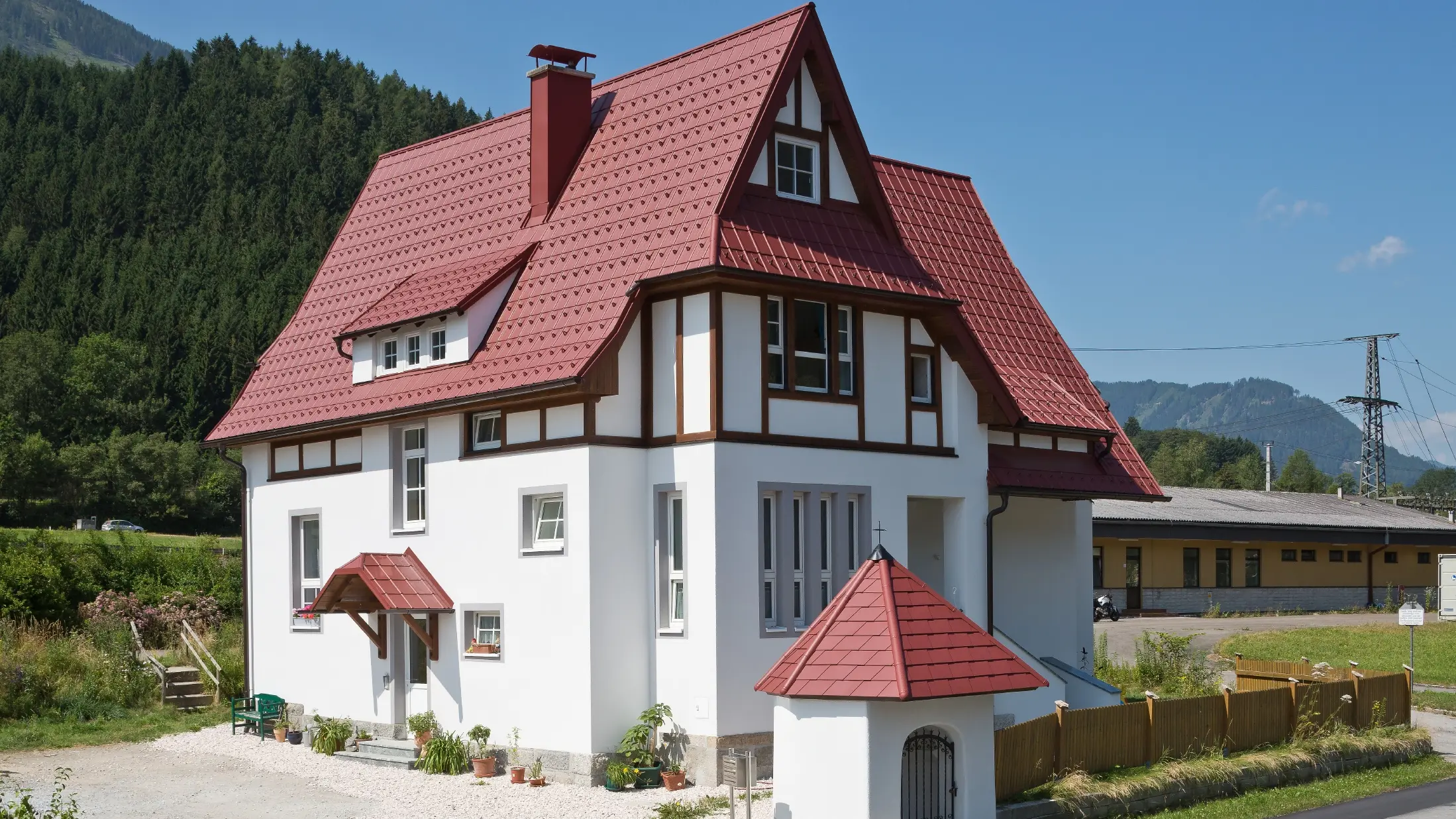 Poševna streha s Strešno ploščo PREFA v oksidno rdeči barvi P.10 s številnimi detajli, kot so strešna okna, stolpiči in dimniški oboki. Tudi nadstrešek in svetišče pred hišo sta pokrita s pločevinasto kritino PREFA.