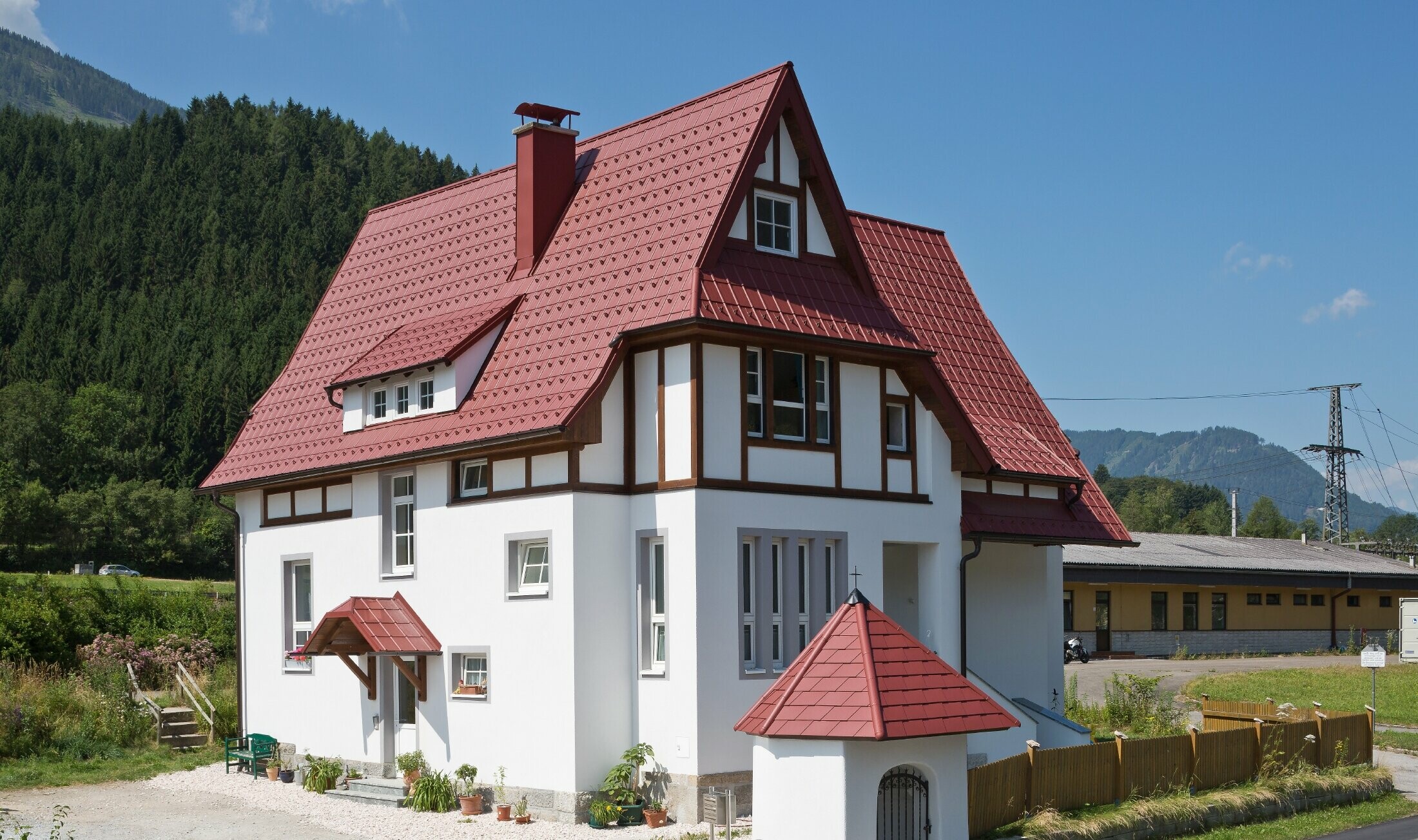 Poševna streha s Strešno ploščo PREFA v oksidno rdeči barvi P.10 s številnimi detajli, kot so strešna okna, stolpiči in dimniški oboki. Tudi nadstrešek in svetišče pred hišo sta pokrita s pločevinasto kritino PREFA.