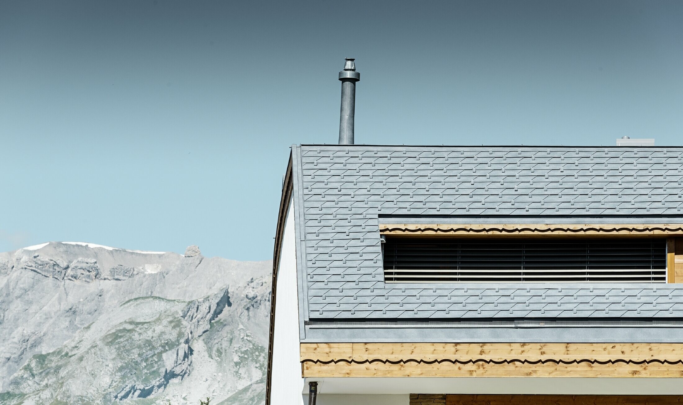 Večdružinska hiša v Crans-Montani z gorami v ozadju in fasado z igrivimi lesenimi elementi in streho iz aluminijastih strešnih skodel PREFA v kamnito sivi barvi