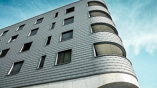 Detajlna fotografija hiše Bernerhaus v Buchsu z izrazito zaobljenim vogalom stavbe in tekočimi balkonskimi odprtinami. Fasada iz PREFA fasadnih skodel v barvi P.10 svetlo siva se kot natančno obdelana aluminijasta koža prilega jasno strukturiranemu volumnu. Luskasta površina poudarja mehke radije in poskrbi za živahno igro svetlobe in sence. Zahvaljujoč lahkemu, vremensko odpornemu in trajnemu PREFA aluminiju nastane homogen ovoj stavbe, ki je enostaven za vzdrževanje – tehnično zahtevna in arhitekturno prava paša za oči.