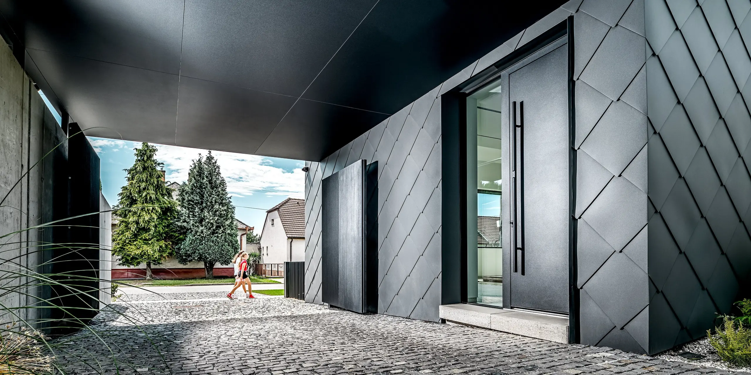 Dieses Bild zeigt den Eingangs- und Durchfahrtsbereich eines modernen Einfamilienhauses in Trnava-Modranka, dessen markante Fassade vollständig mit PREFA Wandrauten 44 × 44 in P.10 Dunkelgrau verkleidet ist. Die präzise Rautenstruktur verleiht dem Gebäude eine elegante, futuristische Wirkung und betont die klare Linienführung der Architektur. Die widerstandsfähigen Aluminiumprodukte sorgen für dauerhaften Schutz vor Witterung und eine hochwertige, langlebige Optik. Im Hintergrund setzt das begrünte Wohnumfeld einen natürlichen Kontrast zur dunklen Gebäudehülle.