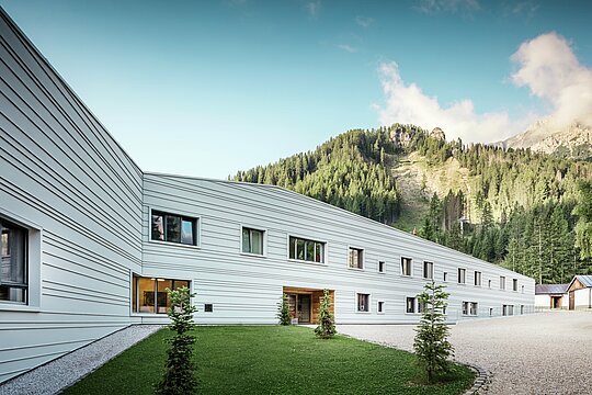 [Translate to Slovenščina:] Aluminium trifft Präzision – ein harmonischer Dialog zwischen Technik und Natur. Vor der eindrucksvollen Kulisse der Dolomiten präsentiert sich das „Villaggio Alpino Tempesti“ mit klarer Linienführung und moderner Gestaltung. Die Fassade aus PREFALZ in der Farbe P.10 Prefaweiß ist nicht nur äußerst widerstandsfähig, sondern sorgt durch ihre helle Farbe auch für ein angenehmes Mikroklima, da sie die Sonnenstrahlen reflektiert. Grünflächen und Holzelemente im Eingangsbereich des Militärgebäudes schaffen eine überraschend einladende Atmosphäre.