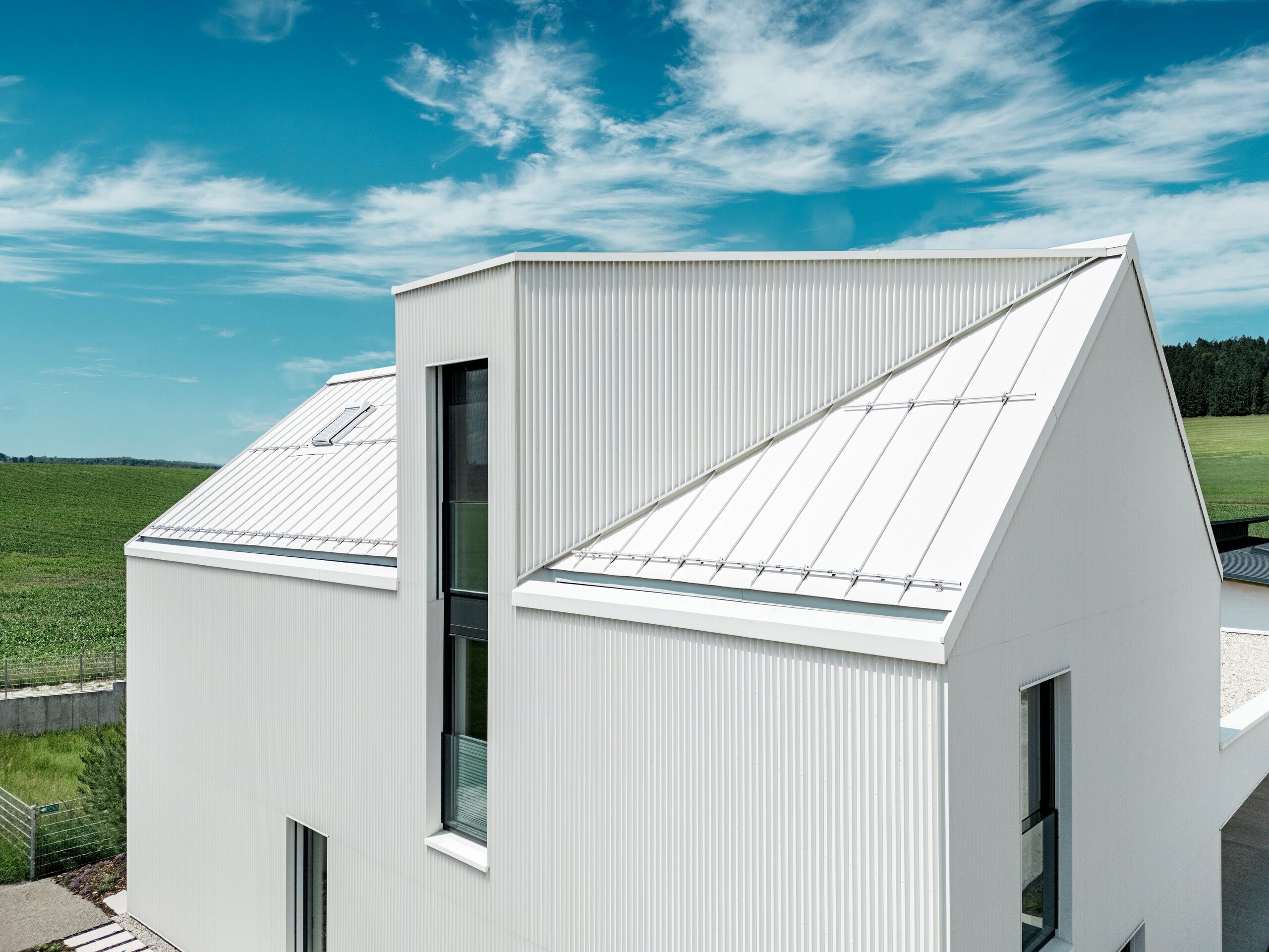 Slika prikazuje strm, izrazit dvokapni strešni pokrov te enodružinske hiše v Freistadtu, ki je obložena s sistemom PREFALZ v beli barvi. Natančno pregibana fasada in streha sta neprekinjeno povezani, kar ustvarja elegantne, jasne linije. PREFALZ, uporabljen za streho in fasado, zagotavlja ne le estetsko trajnost, temveč tudi visoko odpornost proti vremenskim vplivom. To stavbi zagotavlja dolgotrajno trajnost in enostavno vzdrževanje. Sodelovanje sodobnega oblikovanja in funkcionalnih materialov ustvarja harmonično, sodobno arhitekturo, ki je praktična in vizualno privlačna. Posebej v oči padejo strešna okna s poševnim potekom in okni, ki segajo čez dve nadstropji.