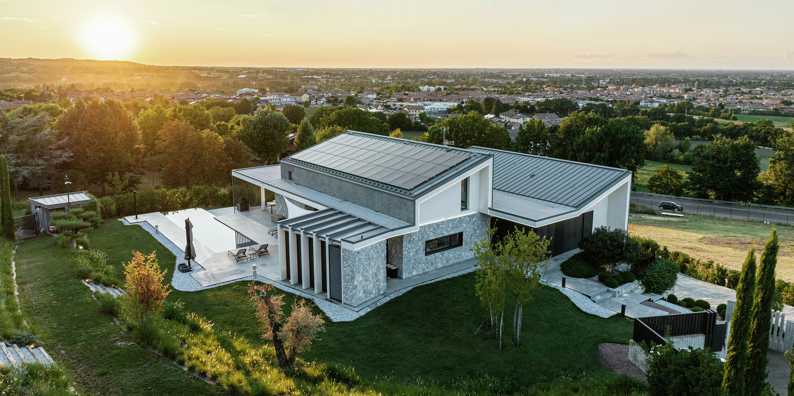 [Translate to Slovenščina:] Das Einfamilienhaus im italienischen Scandiano im warmen Abendlicht. Die beiden Pultdächer mit PREFALZ in P.10 Anthrazit unterstreichen die exklusive Architektur und bieten höchste Langlebigkeit und Widerstandsfähigkeit. Die großzügige Terrasse mit Pool, die harmonische Gartenlandschaft und die moderne Fassadengestaltung runden das luxuriöse Gesamtbild ab. Die großflächige Photovoltaikanlage sorgt für eine umweltfreundliche Energiegewinnung aus Sonnenenergie.