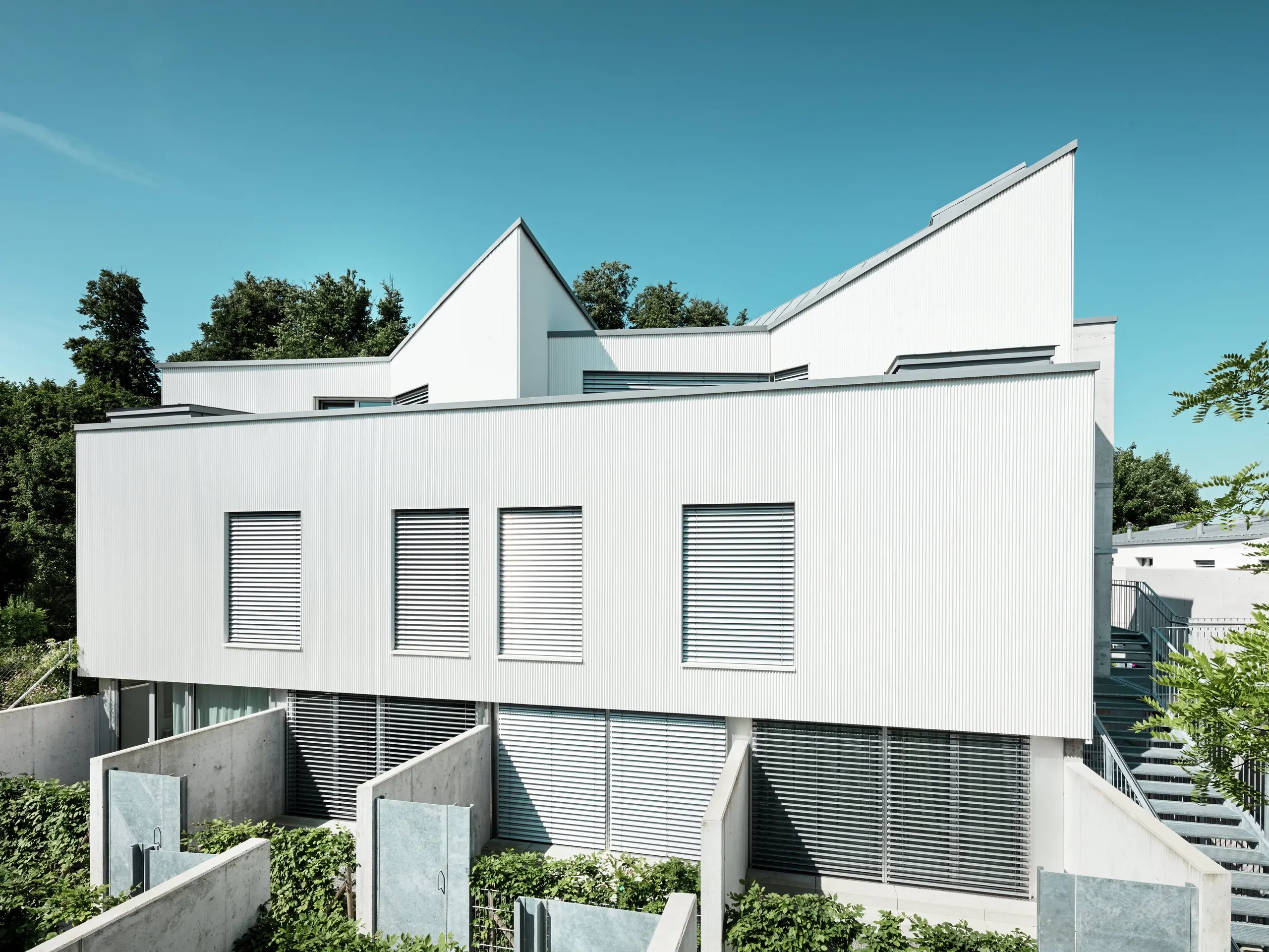 Sodobni Tiny Homes na Zollikerbergu v Švici navdušujejo z jasno linijsko zasnovo in moderno PREFA aluminijasto fasado iz ekstrudirano stikanega zobčastega profila. Navpično vgrajeni, nežno ukrivljeni profili ustvarjajo izrazito in elegantno fasadno podobo, ki jo svetlobni odsevi naredijo živahno. Bela površina daje objektom lahkotnost in poudarja njihovo geometrijsko arhitekturo. Obešena, prezračevana fasada trajno ščiti stavbo pred vremenskimi vplivi ter izboljšuje bivalno klimo. Hkrati je nezahtevna za vzdrževanje in odporna proti koroziji.