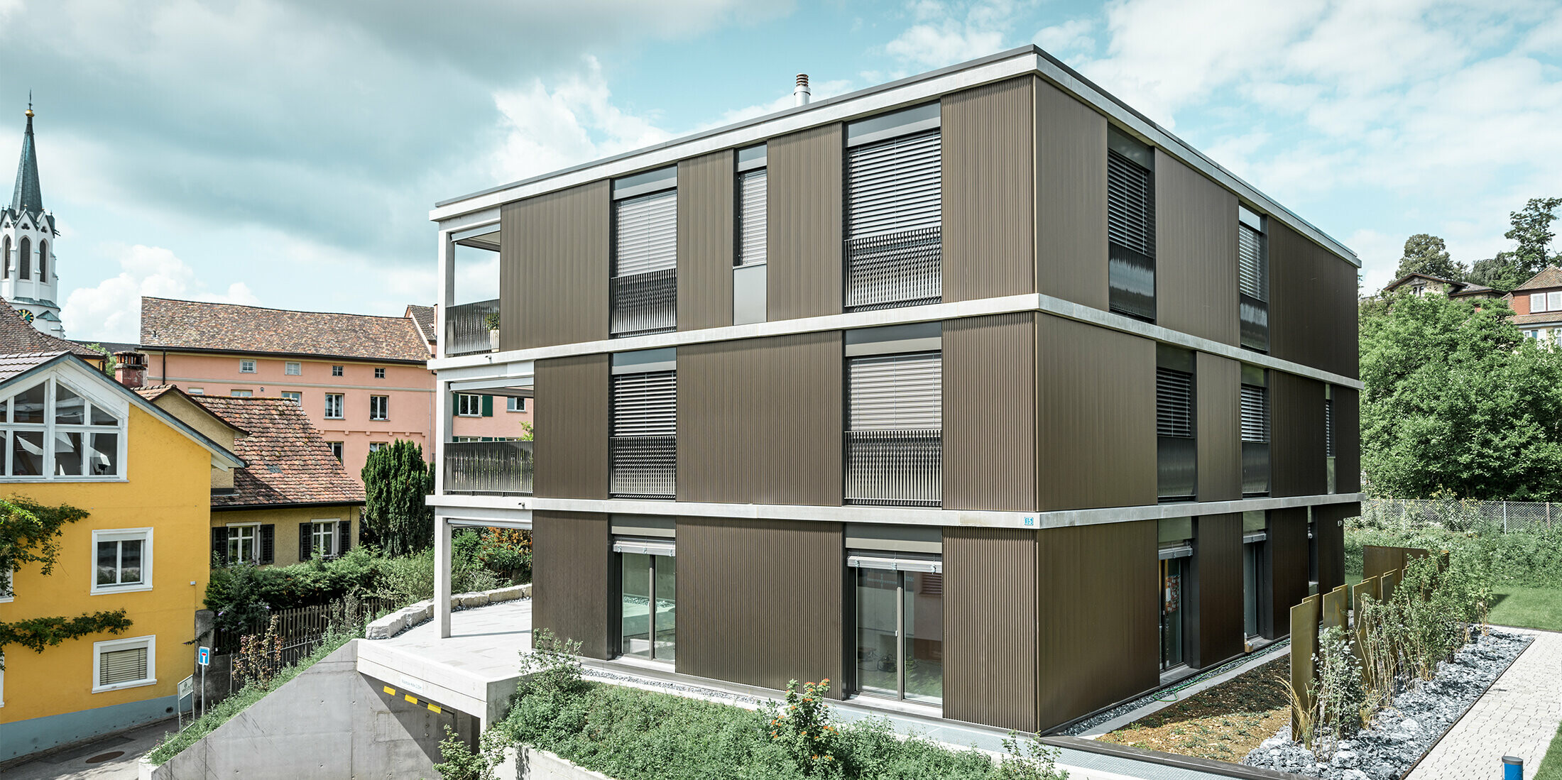 Die markante Aluminiumfassade dieses Mehrfamilienhauses in Schaffhausen ist mit dem eleganten PREFA Zackenprofil in der Farbe Colinal ausgestattet. Durch die vertikale Anordnung der stranggepressten Aluminiumprofile wirkt das Gebäude optisch kompakter und die Fassadenstruktur erhält eine harmonische Balance. Das edle Design fügt sich geschmackvoll in die Umgebung ein und verleiht dem Gebäude eine moderne Eleganz, die sowohl optisch ansprechend als auch funktional ist. Die Perspektive zeigt das Mehrfamilienhaus von der Vorderseite mit der Garageneinfahrt. Im Hintergrund sind Bäume, weitere Gebäude und ein Kirchturm zu erkennen.