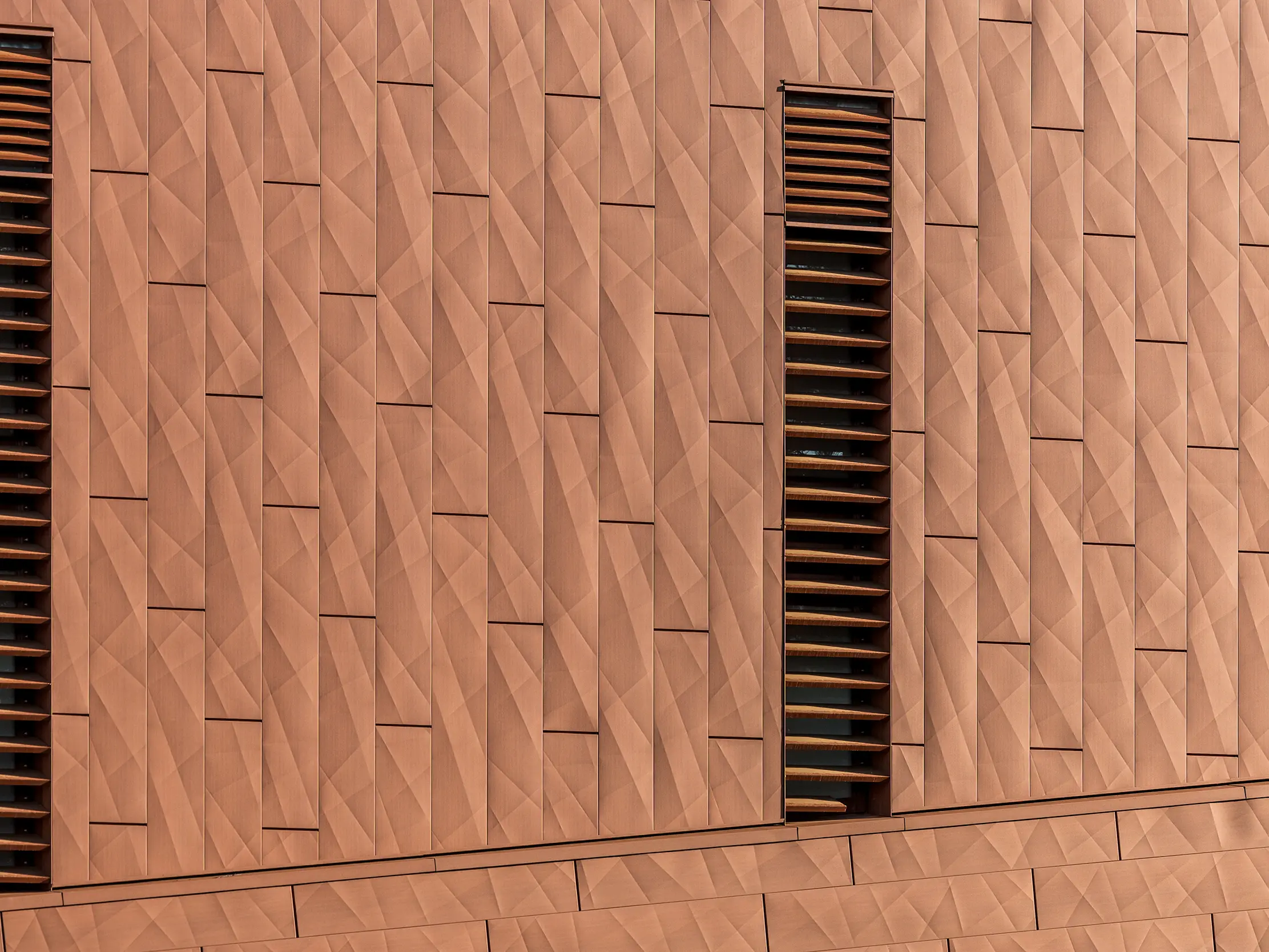 Podrobni posnetek aluminijaste fasade PREFA Siding.X na črpalni postaji pitne vode WML v Lottumu z drobno gubano, geometrično površino v topli rjavi barvi. Natančno razporejeni paneli ustvarjajo živahno igro svetlobe in sence, medtem ko vitke, navpične lamele kot izraziti poudarki strukturirajo videz fasade. Lahki, trajni in v celoti reciklirani PREFA aluminijasti paneli prepričajo z visoko odpornostjo proti vremenskim vplivom, barvno obstojnostjo ter trajnostno in vzdrževanju prijazno ovojnico stavbe.
