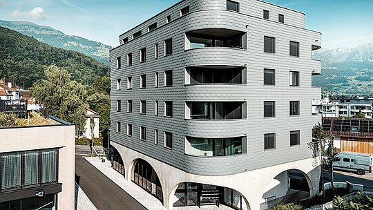 Zunanji pogled na hišo Bernerhaus v Buchsu z značilno trikotno obliko in nežno zaobljenimi balkonskimi pasovi. Fasada iz PREFA fasadnih skodel v barvi P.10 svetlo siva obdaja stavbo kot luskasta aluminijasta koža in ustvarja natančen, živahen videz fasade. Nad masivnim betonskim podstavkom z elegantnimi arkadami deluje lahka, vremensko odporna PREFA aluminijasta fasada še posebej izrazito. Trajna, enostavna za vzdrževanje in oblikovno stabilna postavlja pred alpskim gorskim ozadjem sodoben arhitekturni poudarek.