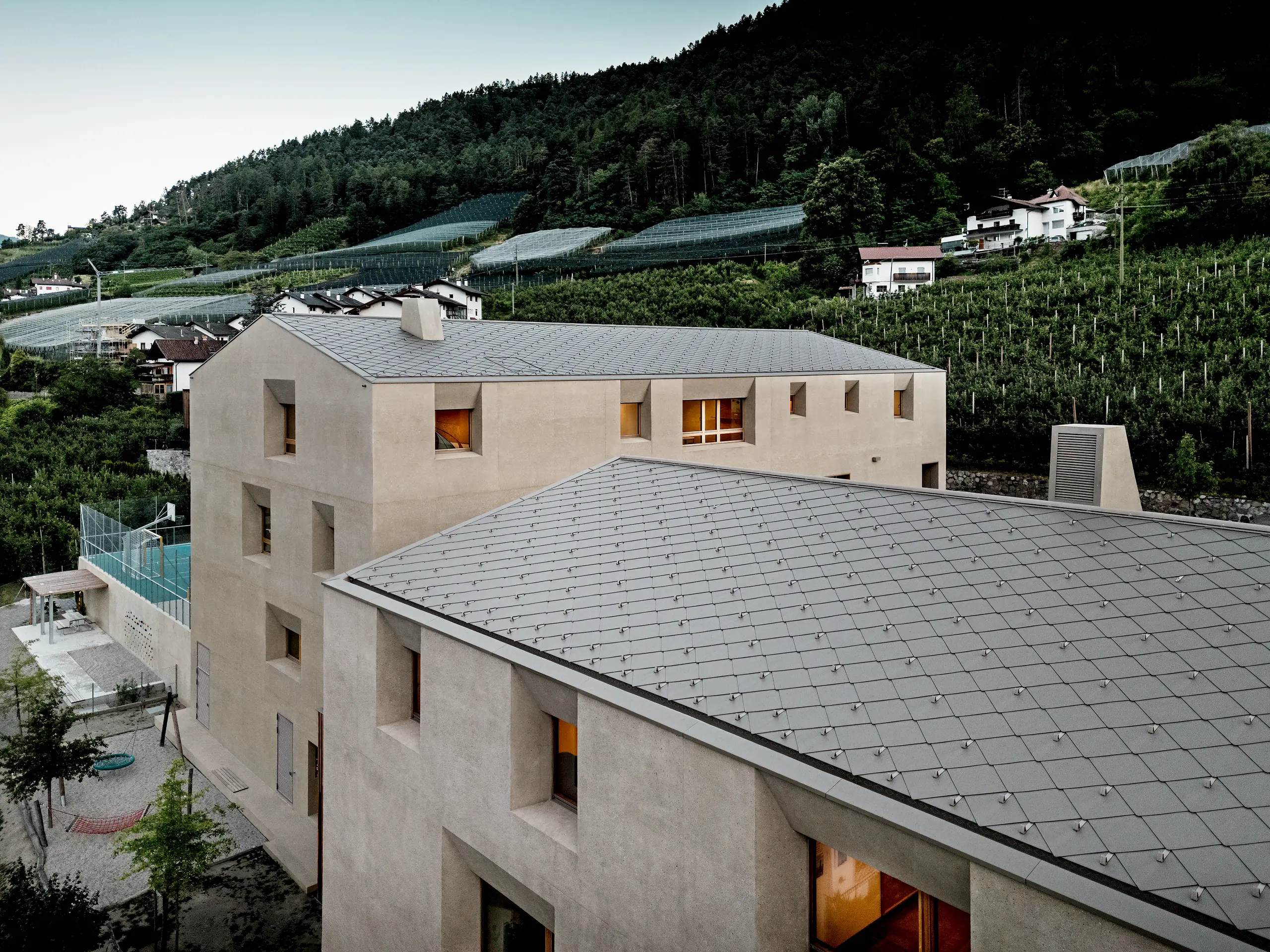 Iz ptičje perspektive izobraževalni center Riffian na Južnem Tirolskem razkriva svoje markantne strešne rombe PREFA 44 × 44 v barvi P.10 bronasta. Natančno izvedena aluminijasta strešna kritina elegantno prekriva vrtec in osnovno šolo, zagotavlja dolgotrajno zaščito pred vremenskimi vplivi ter omogoča, da se sodobna arhitektura harmonično zliva z okoliškimi sadovnjaki in gorskim pobočjem.