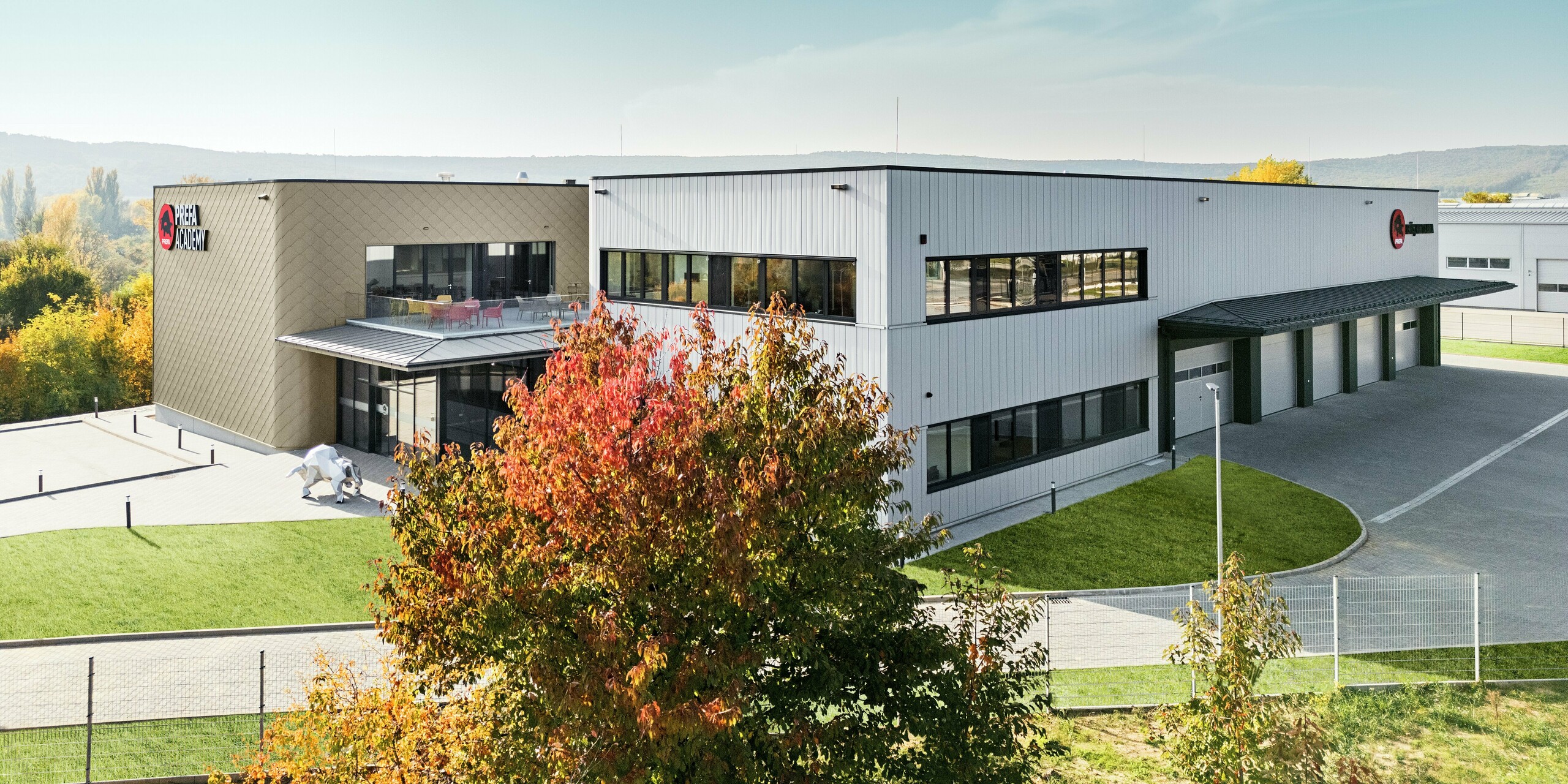 Nova lokacija PREFA Madžarska v Törökbálintu navdušuje z moderno fasadno zasnovo iz aluminijastih rombov PREFA 44 × 44 v bronasti barvi in fasadnih elementov PREFA Siding v srebrno kovinski barvi. Kombinacija zagotavlja elegantno in hkrati robustno zunanjo ovojnico, ki je trpežna in odporna na vremenske vplive. Prostorna stavba združuje učilnice, pisarne ter obsežno skladišče in hkrati predstavlja arhitekturni izraz inovativnosti in kakovosti.
