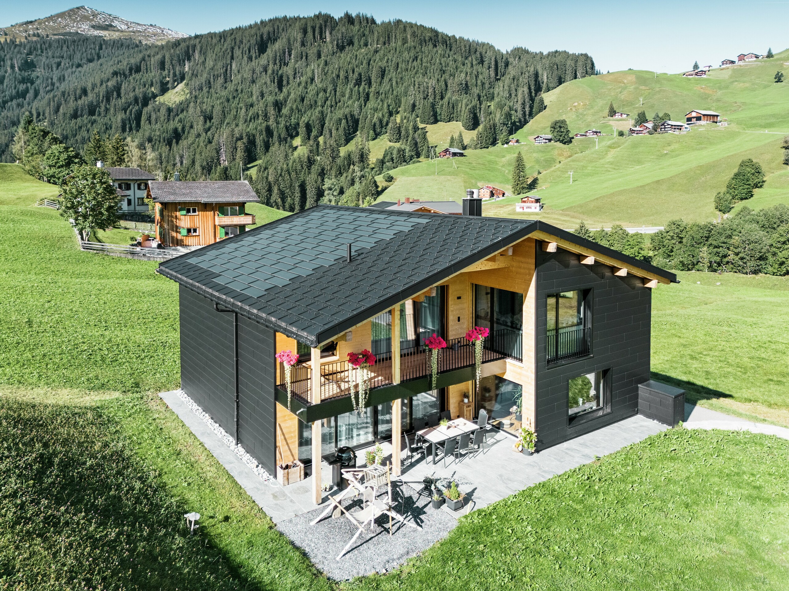 Elegantna enodružinska hiša v hribovitem okolju Ascharina (Švica) kaže, kako se lahko aluminij in les odlično združita. Robustni fasadni paneli PREFA Siding.X v črni barvi P.10 so usklajeni s streho iz solarnih strešnih plošč PREFA in strešnih plošč R.16 v enakem barvnem premazu. Trpežna aluminijasta obloga zanesljivo ščiti stavbo pred izzivi alpskega podnebja.